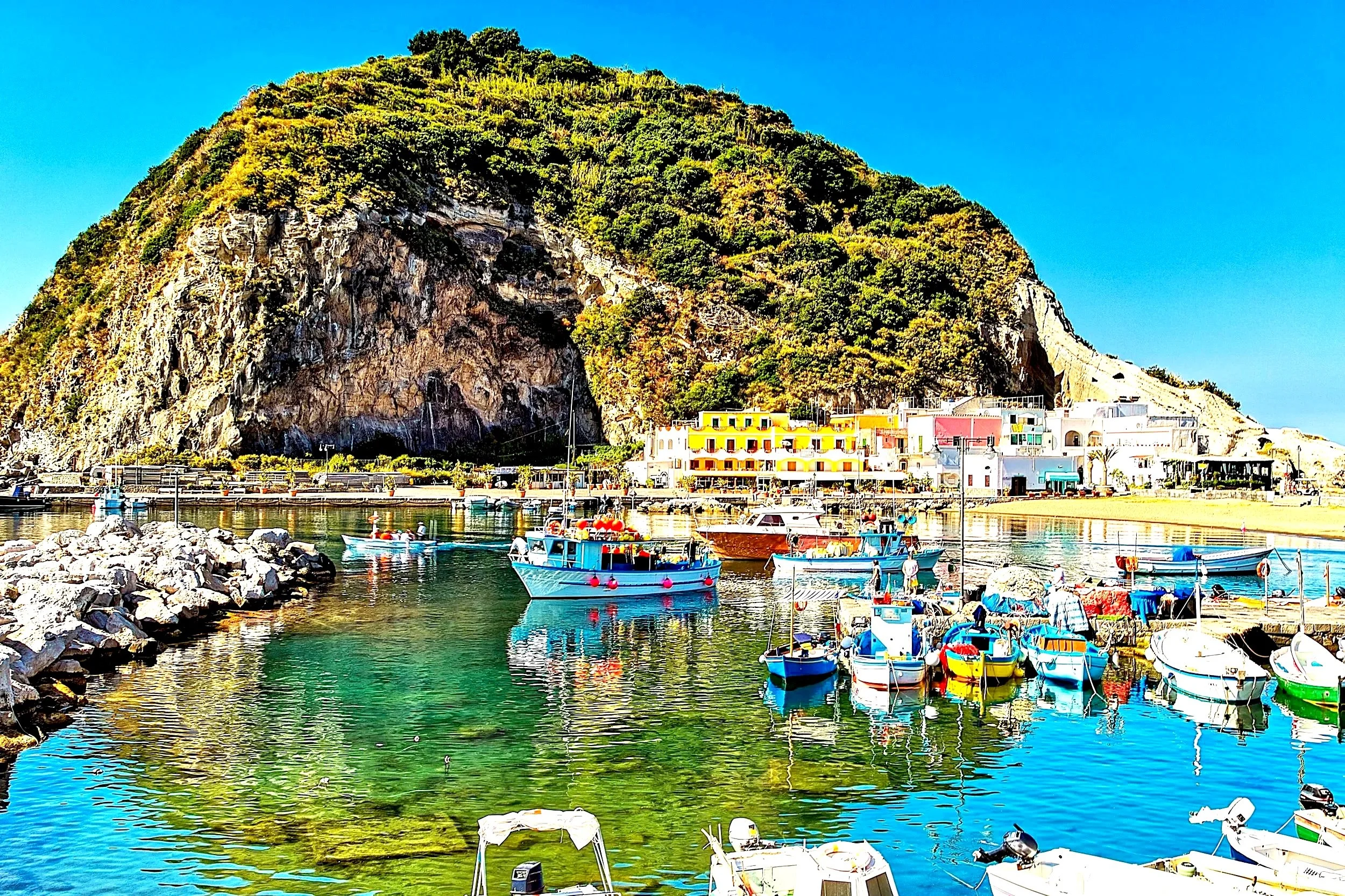 Colorful boats docked in a small harbor with a hillside town and green mountain in the background.