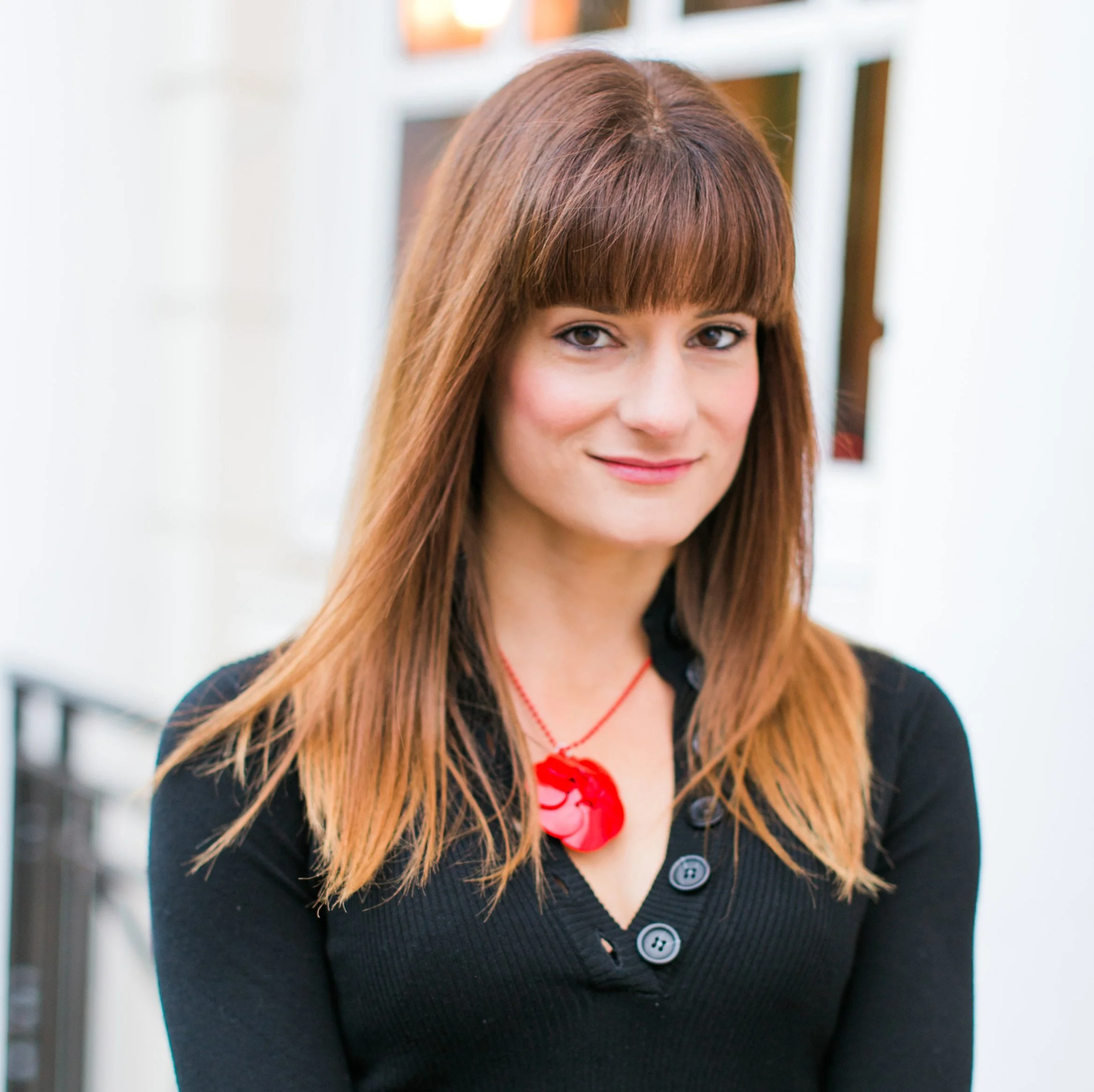 A woman with long brown hair and bangs, wearing a black button-up top and a red necklace, standing outdoors with a white building and a black railing in the background.