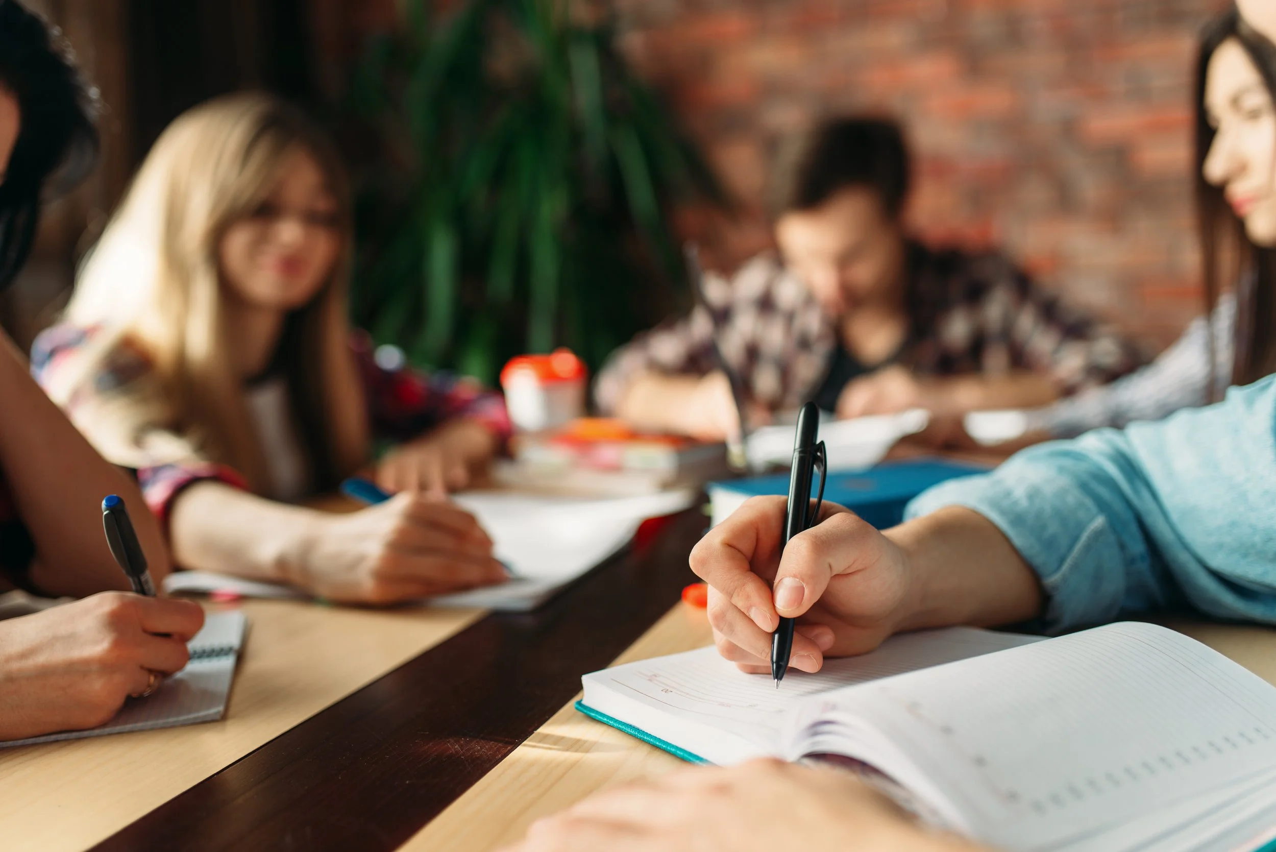 People sitting at a table, writing in notebooks, with a brick wall and indoor plants in the background.