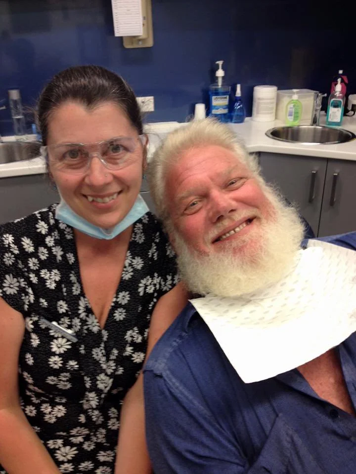 A woman with glasses and a face mask around her neck smiling next to an older man with a white beard, both in a medical or dental office environment.