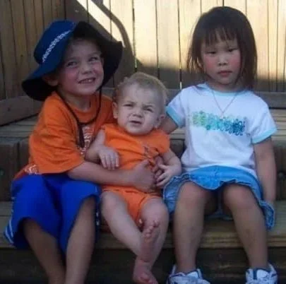 Three children sitting on a wooden bench in a backyard, two girls and a boy, with a wooden fence in the background.