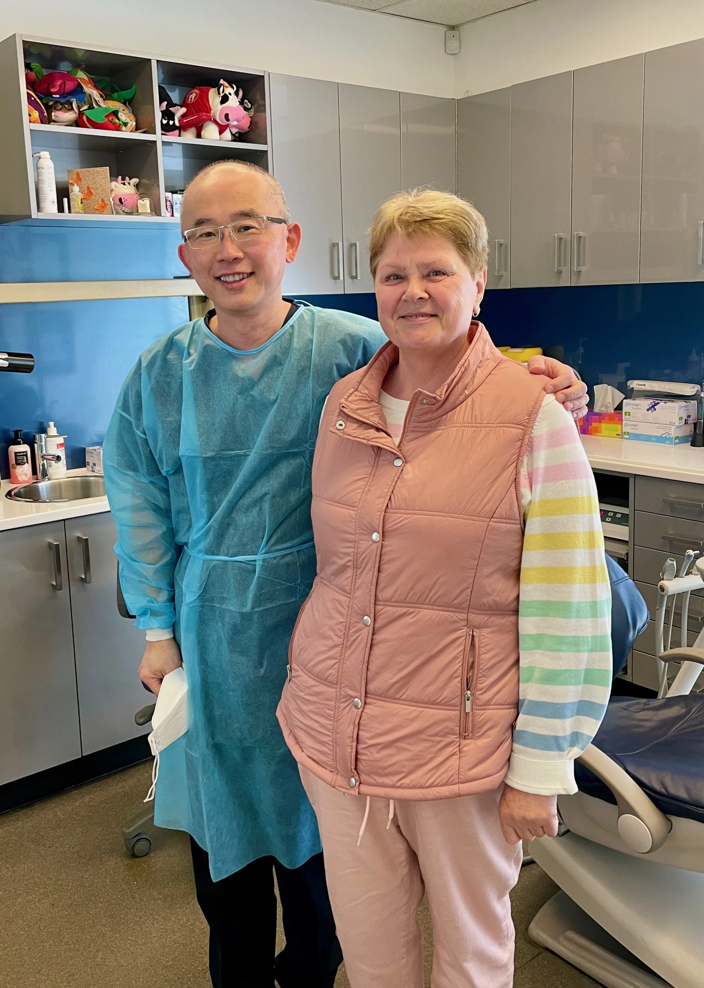 A healthcare worker and an elderly woman smiling in a medical clinic, with medical supplies and colorful toys on shelves in the background.