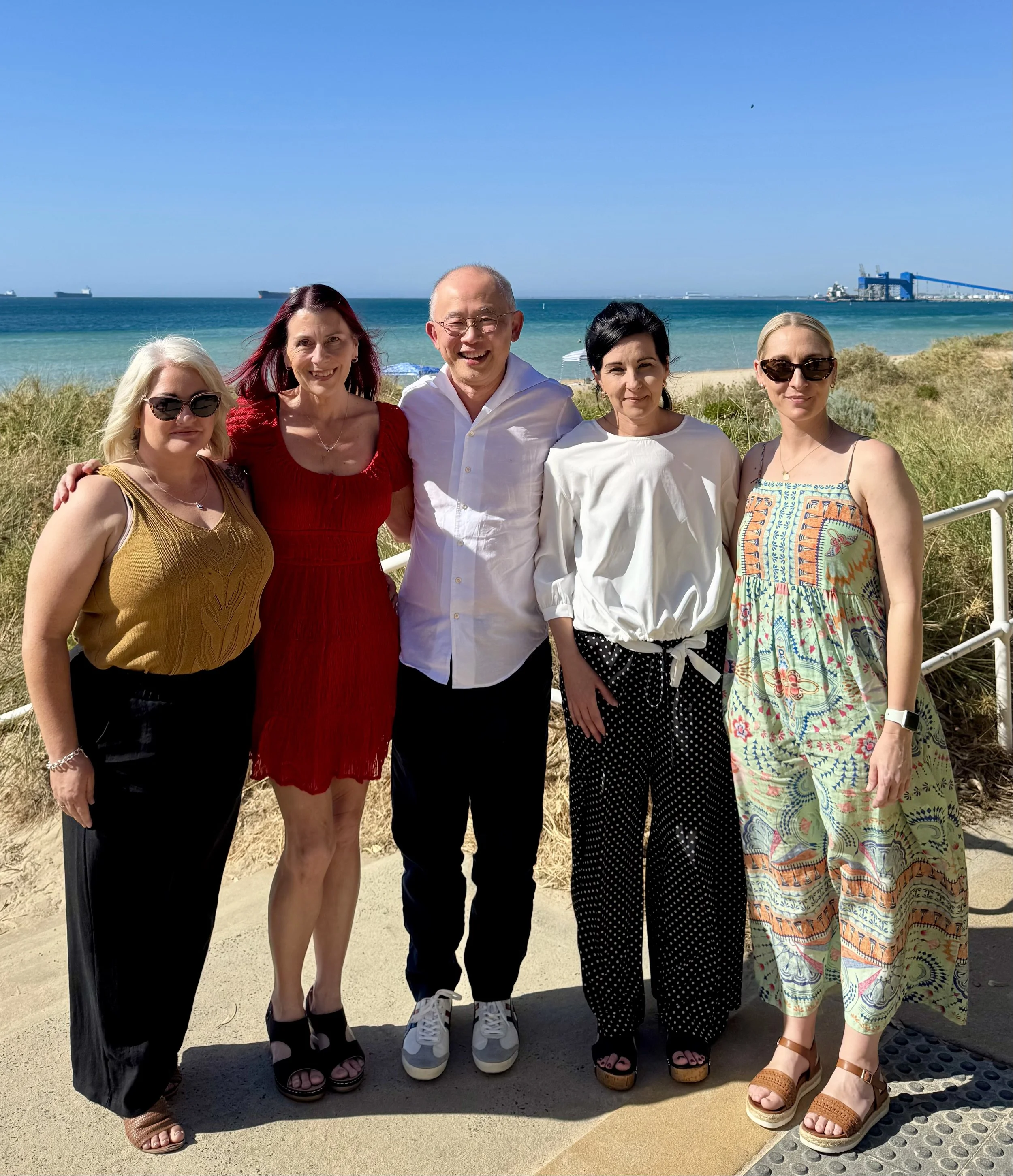 Five women and one man standing together on a beach path with the ocean and ships in the background, smiling for the photo.