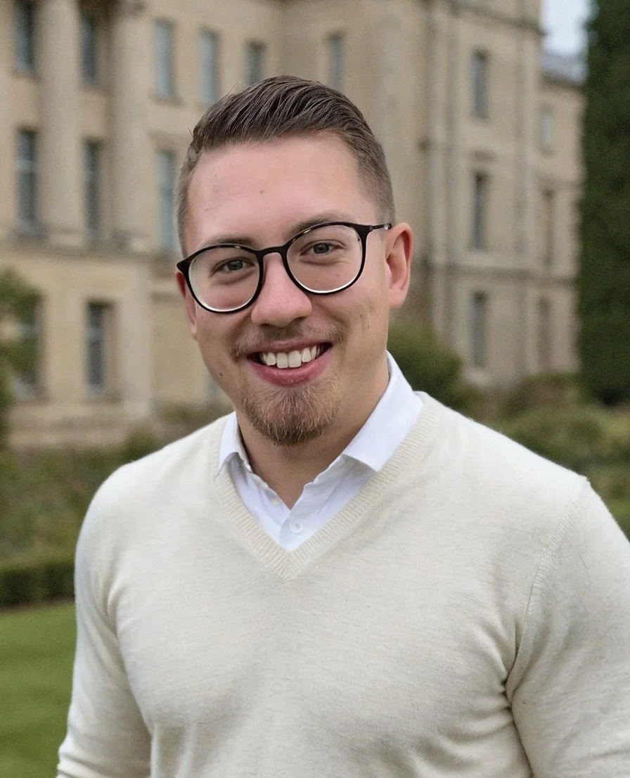 Junger Mann mit Brille, Lächeln, hellen Haaren, trägt weißes Hemd und hellgelben Pullover, im Hintergrund Graste und ein altes Gebäude.