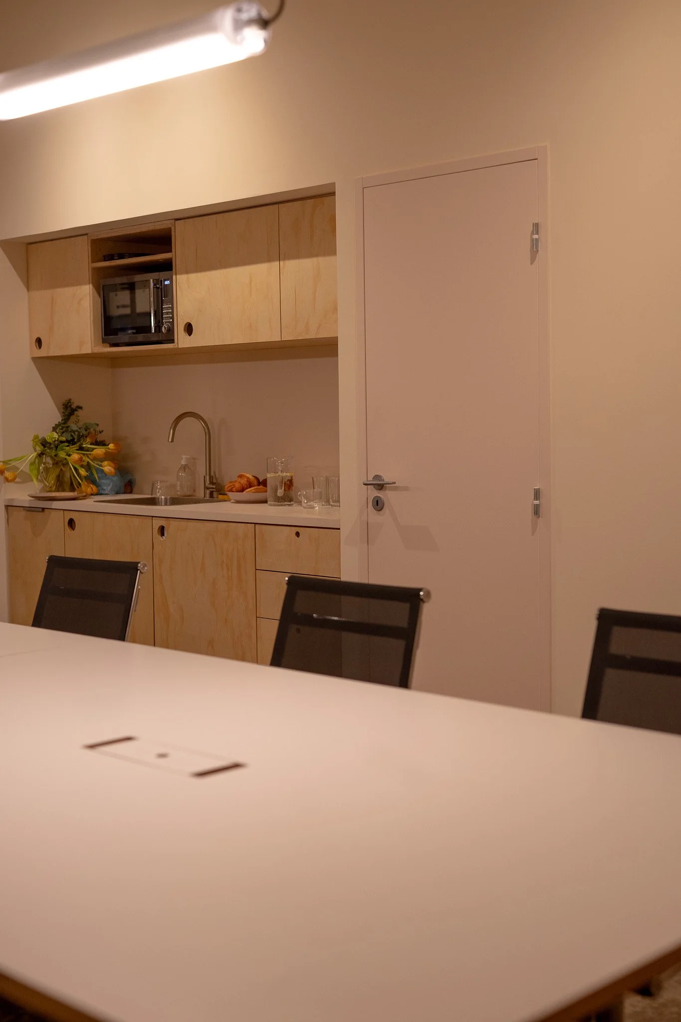 Openspace, chaises noires, petite cuisine, four micro-ondes, plantes et petits objets décoratifs, mur beige et porte blanche.