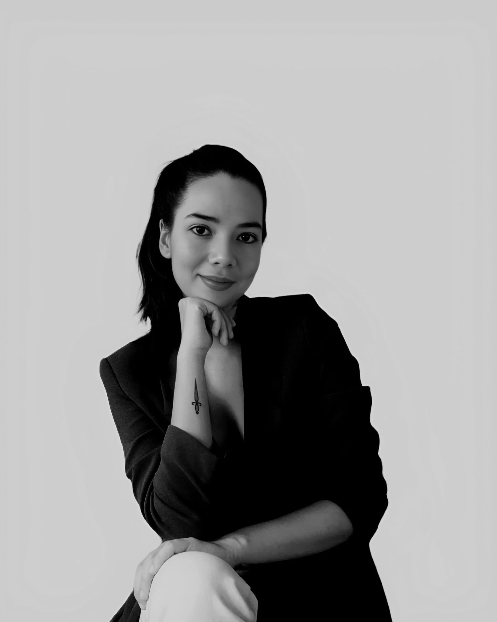 Black and white portrait of a woman with dark hair, smiling gently, wearing a dark blazer with her right hand resting on her chin, and a tattoo of a small sword on her forearm.
