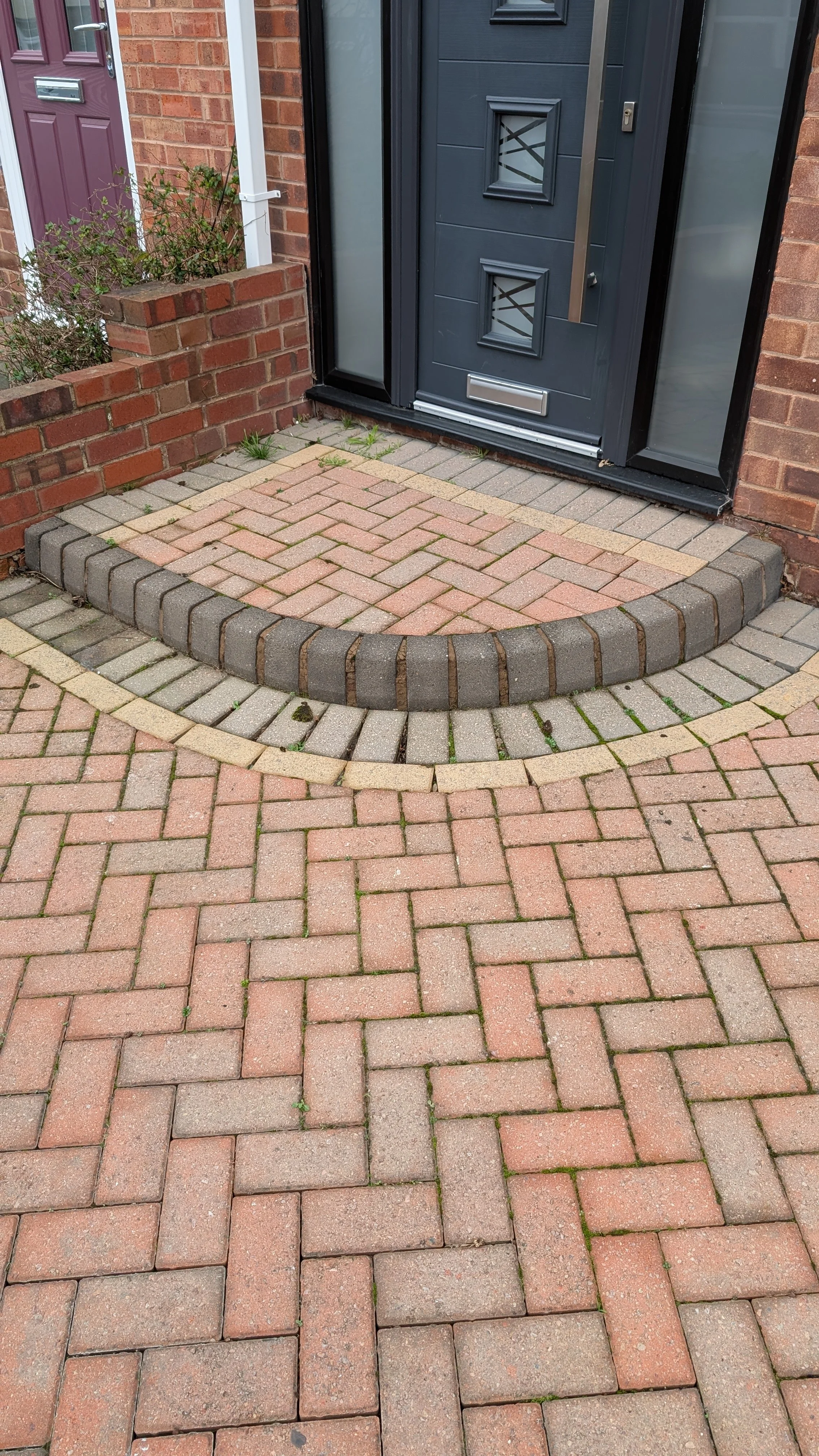 Close-up of a brick front porch with a black door and a small garden bed to the left, surrounded by brick walls and a brick walkway.