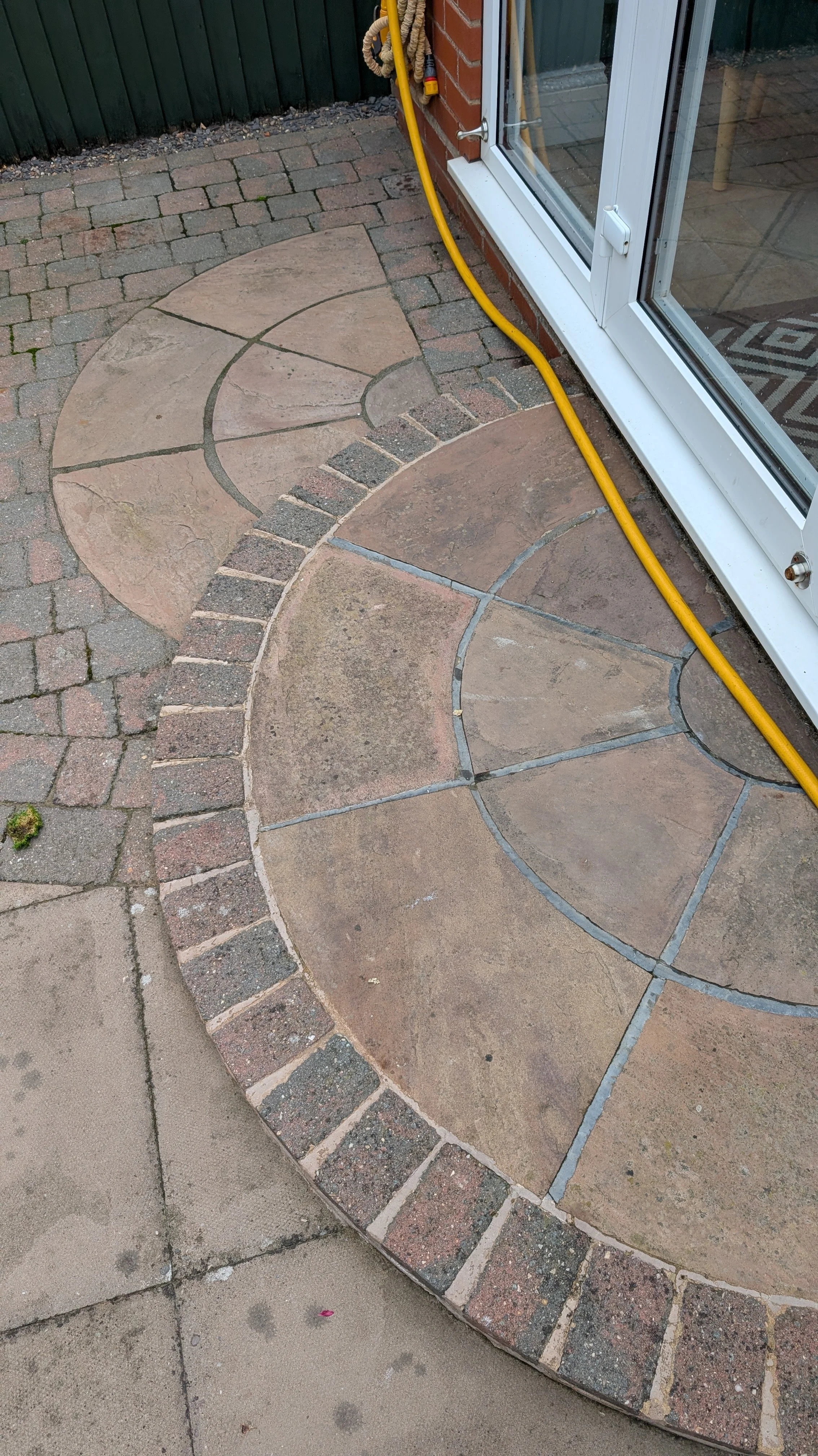 Decorative outdoor patio with circular and semi-circular stone and brick pattern, adjacent to a glass door and a yellow hose.