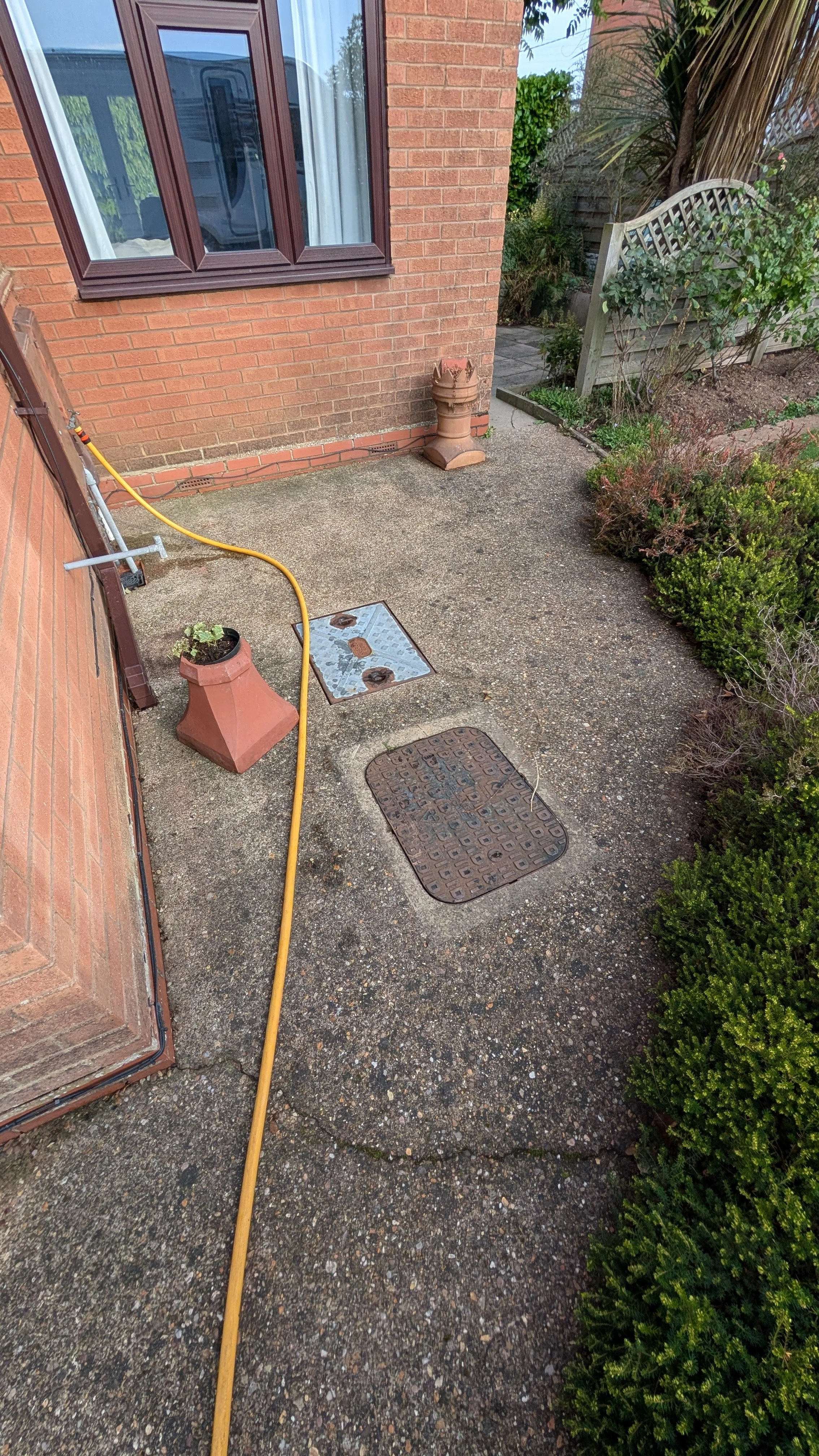 A backyard patio with a concrete surface, a brick house wall, a window, a garden hose, a potted plant, a decorative planter, and some bushes and trees.