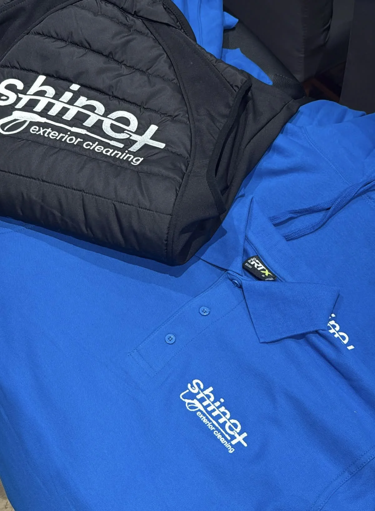 A black bag with the logo 'Shine Shinet exterior cleaning' and a blue polo shirt with the same logo laid on a surface.