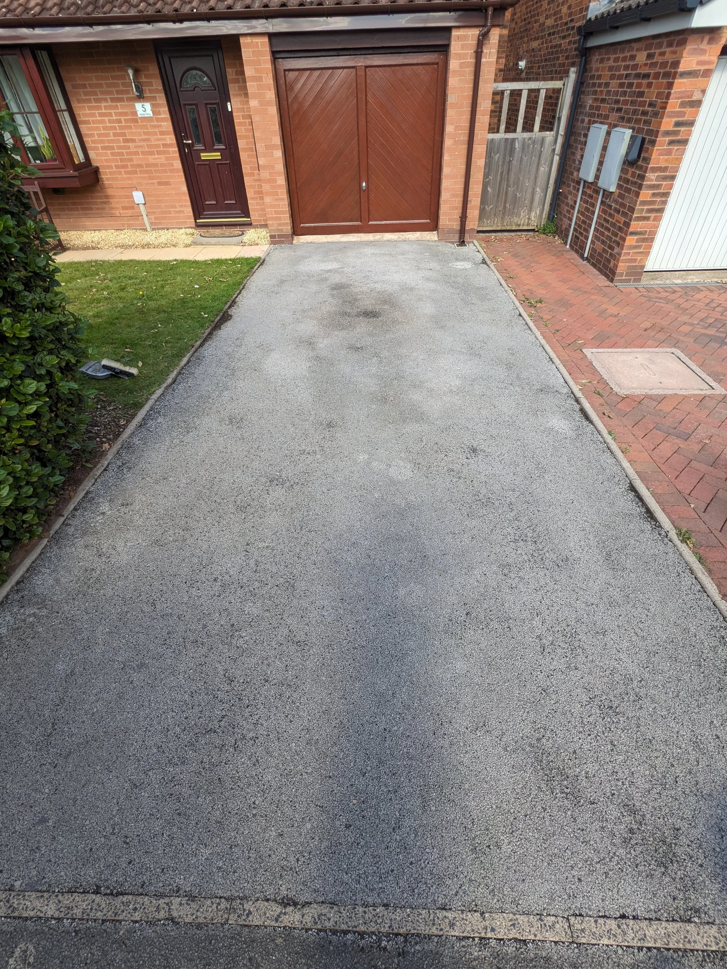 Residential driveway made of gravel with a house in the background featuring a dark wooden door, a garage, and brick walls. To the left is a small manicured lawn with a bush, and to the right is a brick sidewalk with utility meters.
