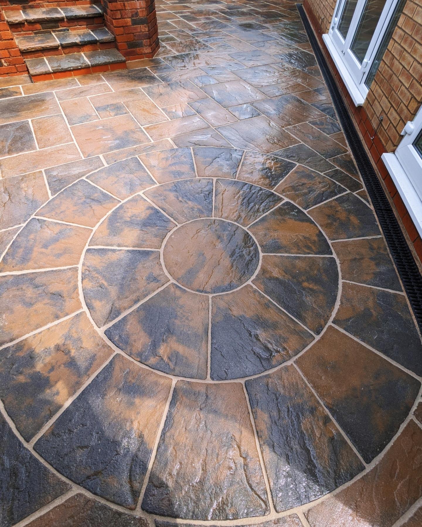 A decorative slate tile patio with a circular pattern in the center, adjacent to a brick house with large windows and steps leading to another area.