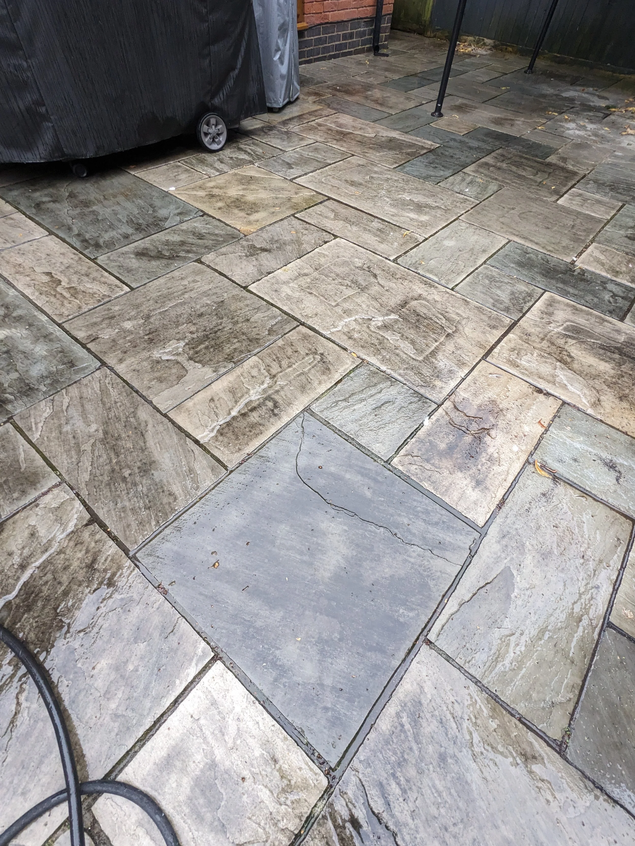 Stone patio with variously sized rectangular and square tiles in neutral tones, with some leaves and a black cable in the foreground, and a black trash bin and outdoor furniture in the background.