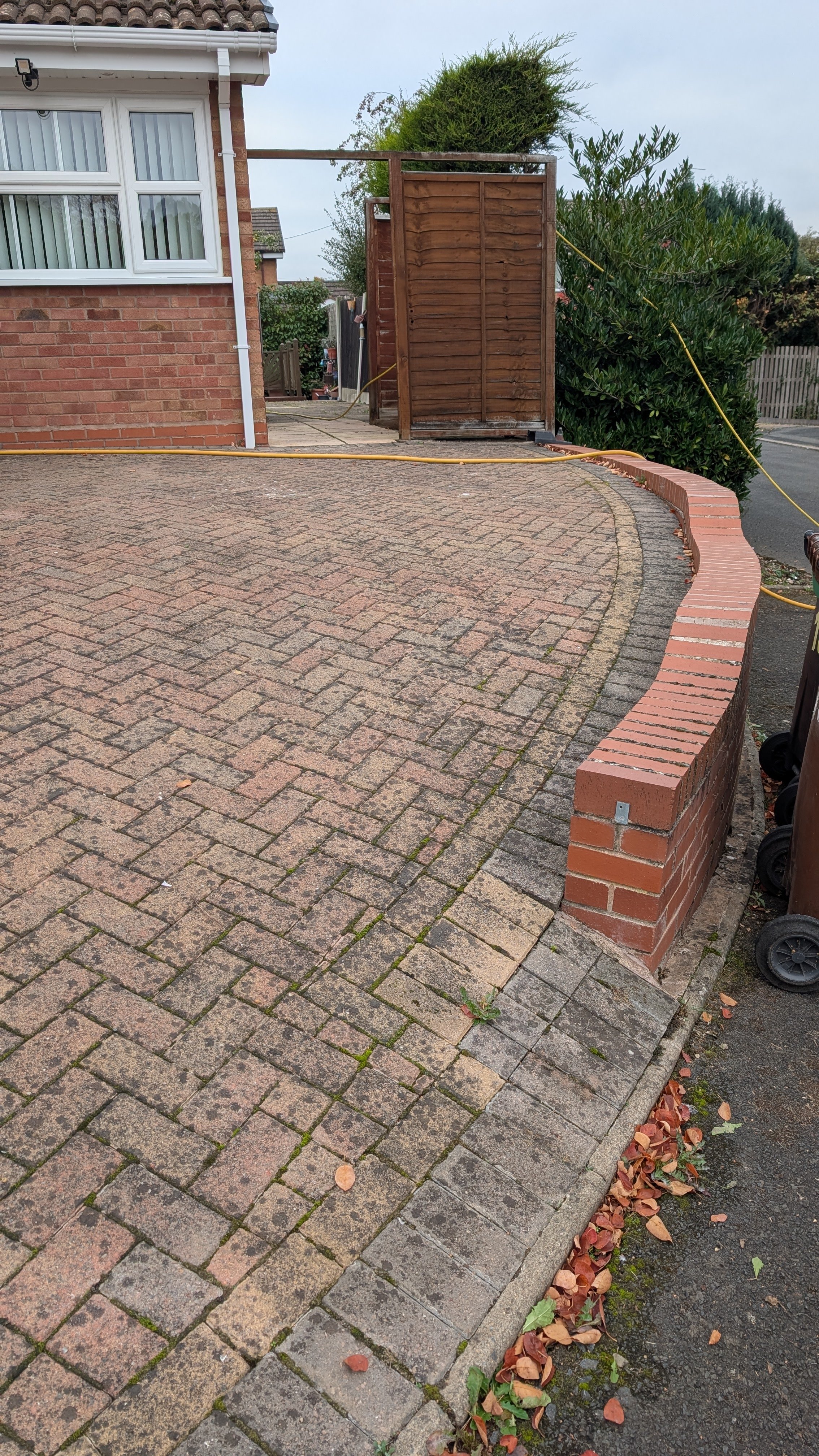 View of a brick driveway with a curved brick retaining wall on the right side, a garden gate at the top of the driveway, and some fallen leaves along the edge of the driveway.