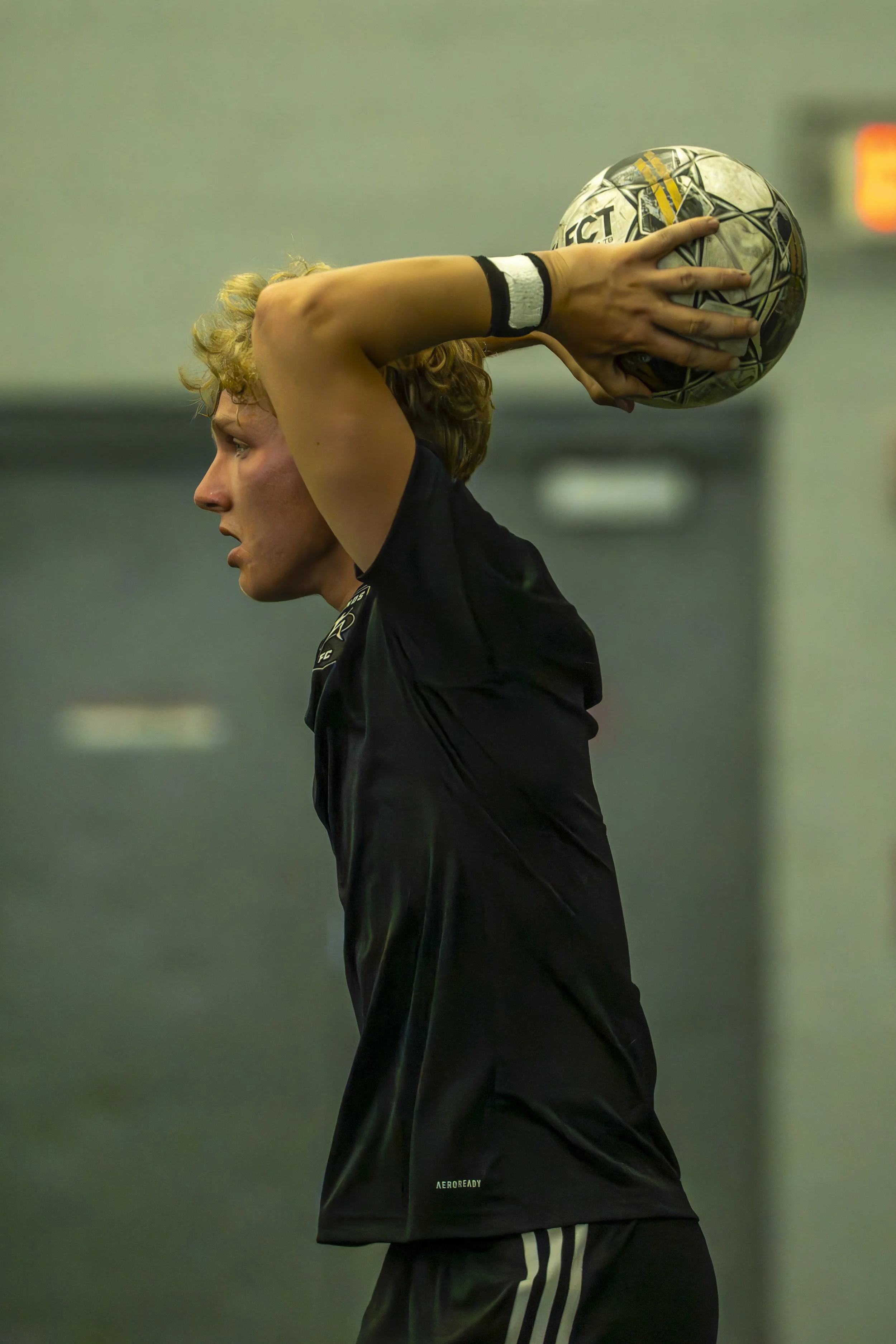 A female athlete with curly blond hair in a black sports uniform holding a soccer ball above her head during a game or practice indoors.