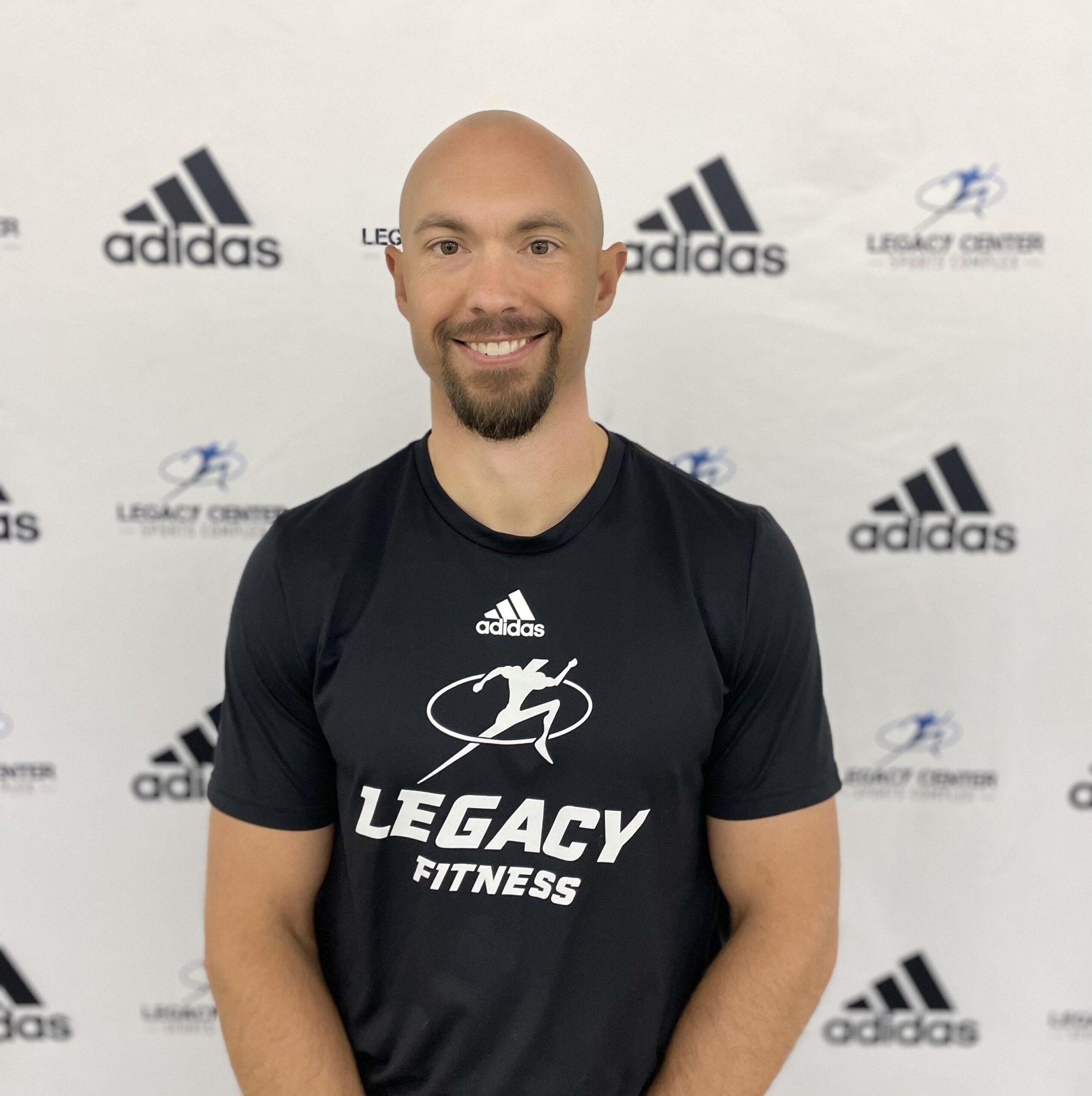 A man smiling in front of a white background with Adidas and Legacy Center logos, wearing a black T-shirt with the Legacy Fitness logo.