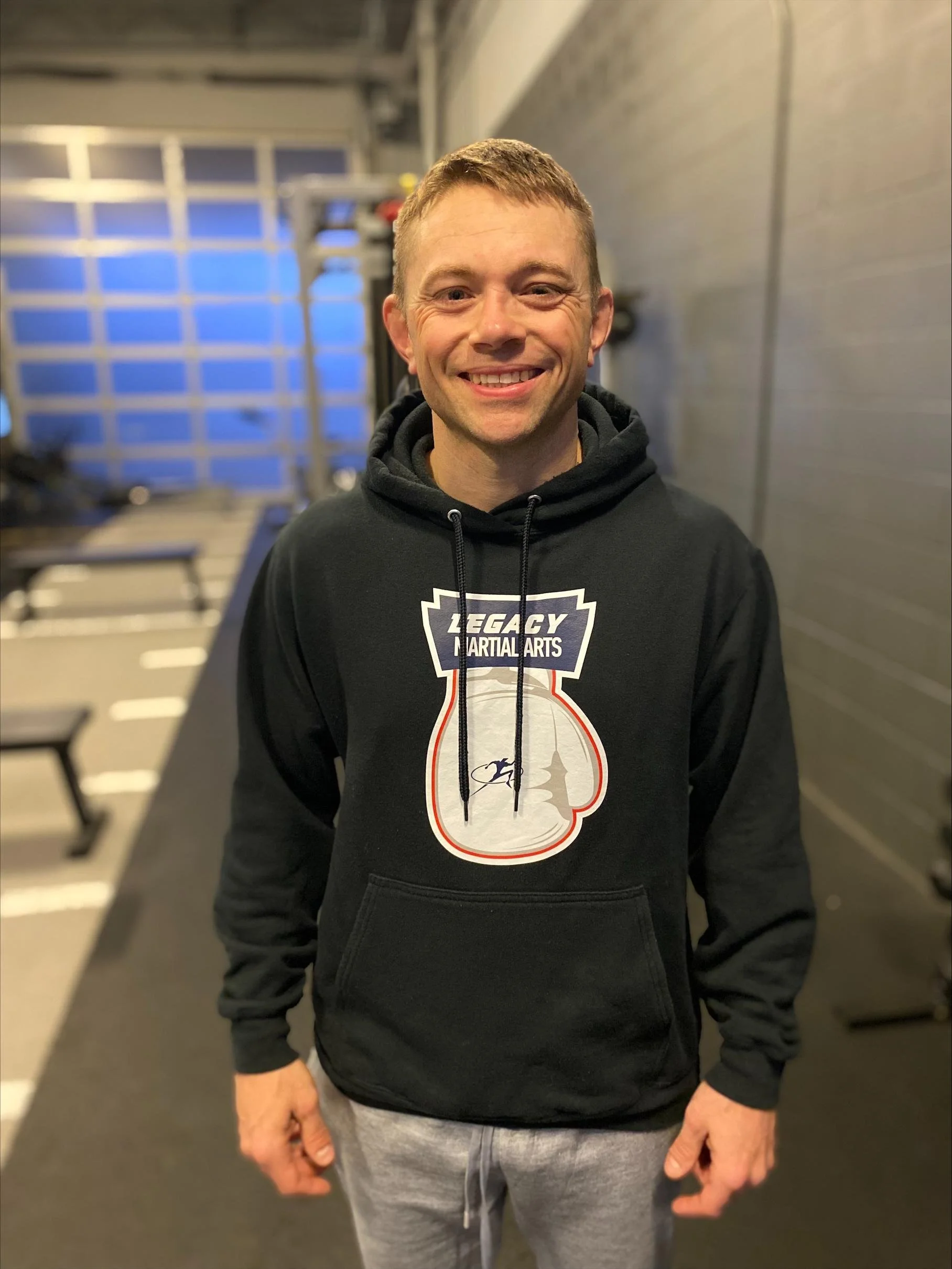 A smiling man in a black hoodie with 'Legacy Martial Arts' logo, standing inside a gym with weightlifting equipment.