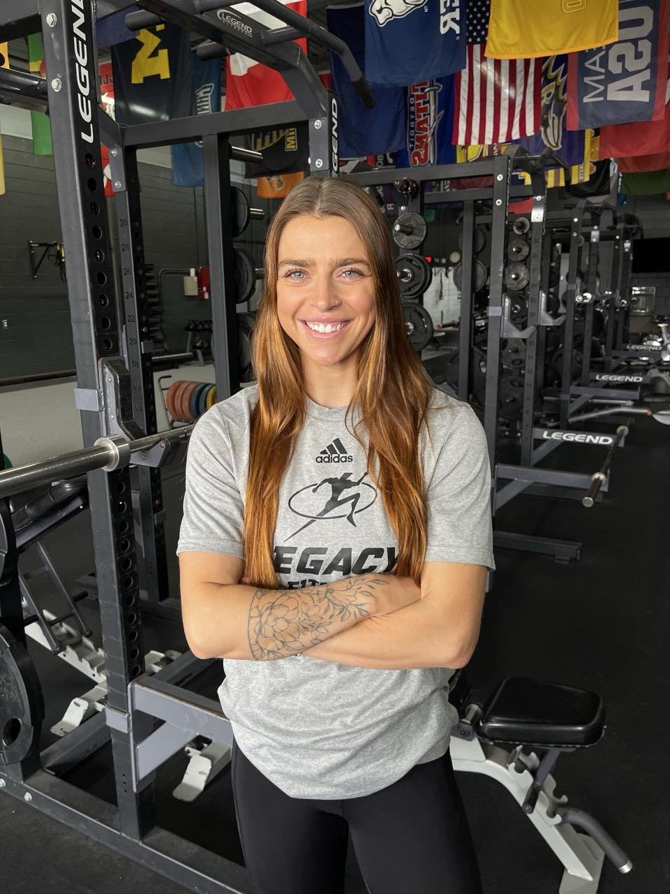 A woman smiling with arms crossed in a gym, surrounded by weightlifting equipment and American flags hanging from the ceiling.