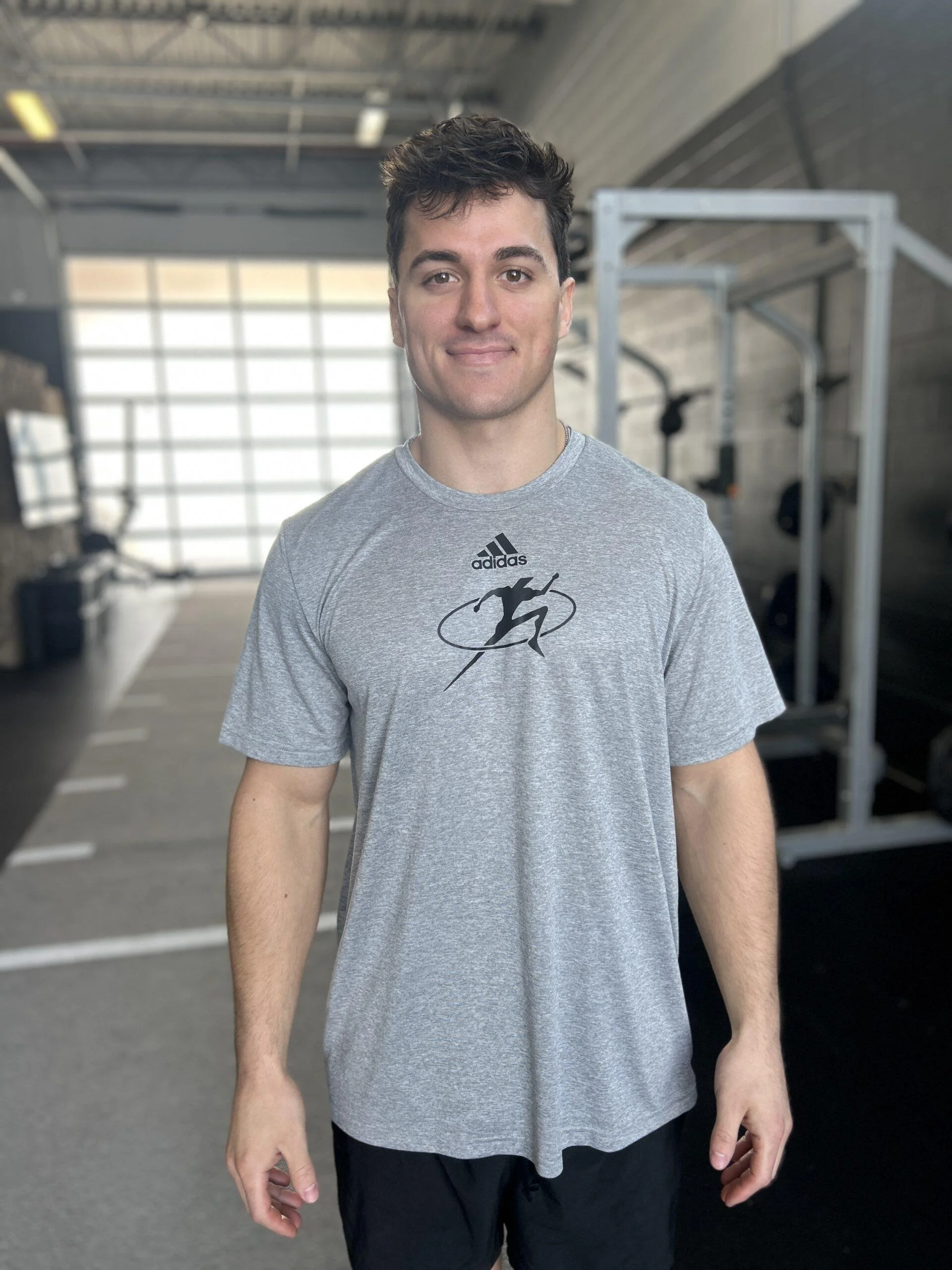 A young man with short, curly brown hair, wearing a gray athletic t-shirt with a black running figure logo and black shorts, standing in a gym.
