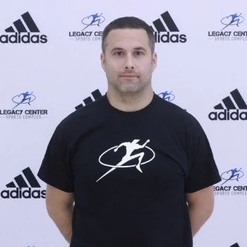 A man standing in front of a backdrop with Adidas and Legacy Center logos, wearing a black t-shirt with a white logo.