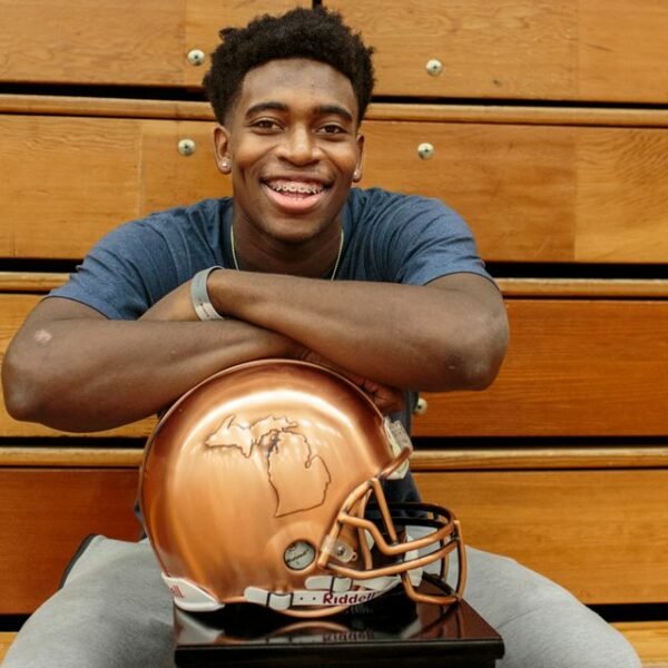 A young man with short curly hair and braces smiling at the camera, sitting behind a football helmet with a Michigan map logo, on a wooden background.