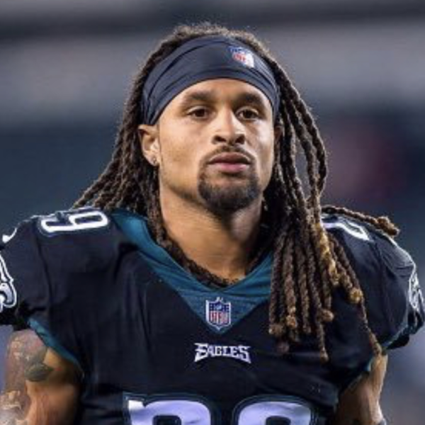 A football player in a black and teal jersey with a headband, shoulder pads, and long dreadlocks.