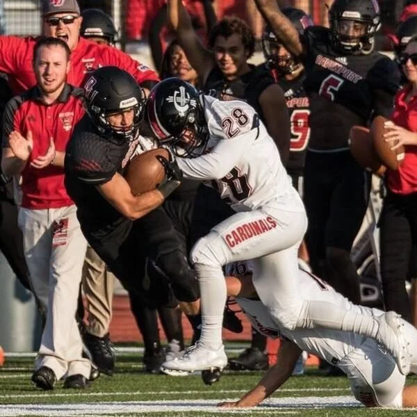 Football players in action during a game, with one player carrying the ball and another tackling him, while teammates and spectators watch.