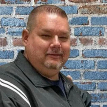 A man with short hair and a goatee, wearing a dark jacket, standing in front of a blue brick wall.