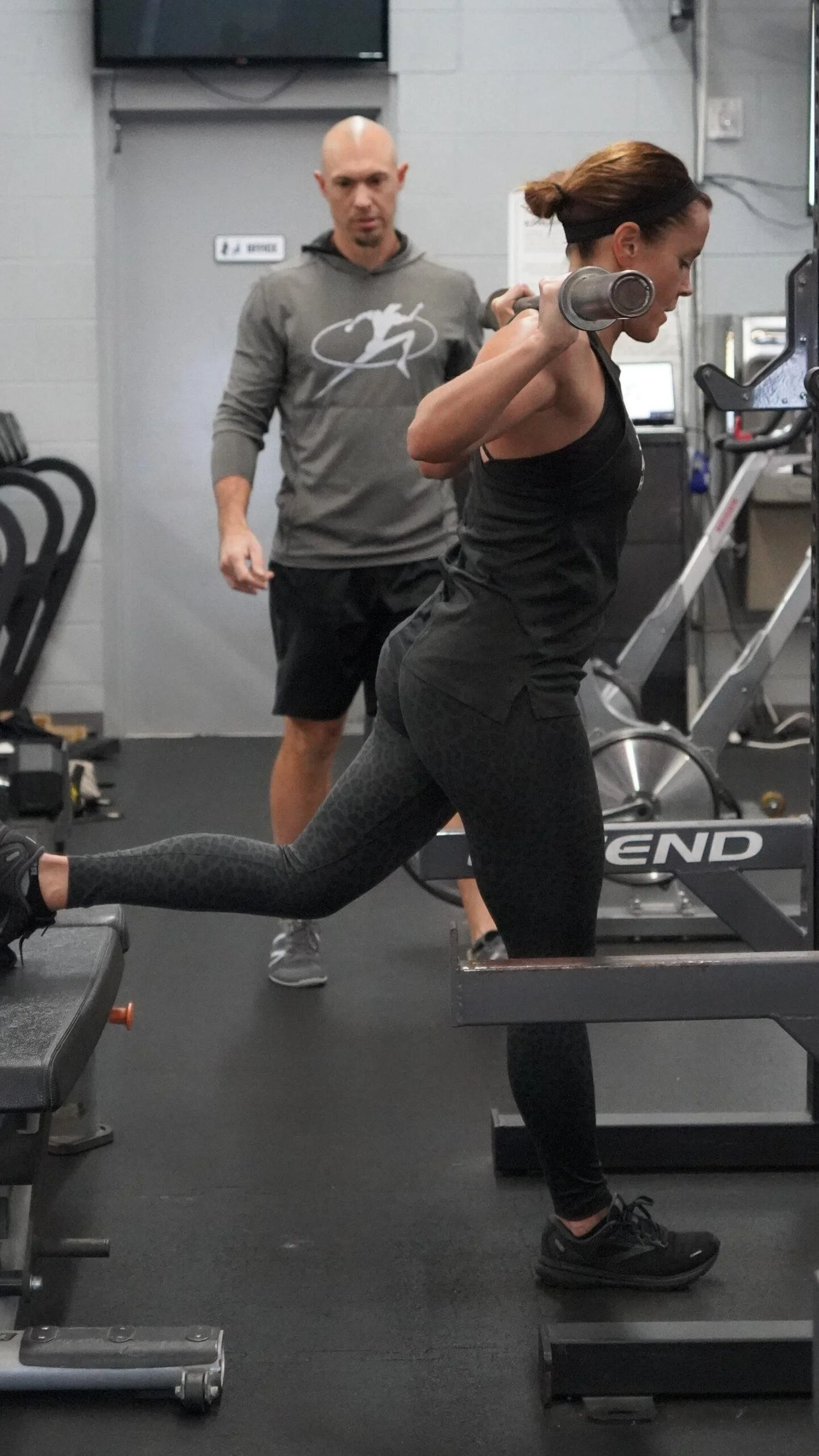 A woman performs a step-up exercise holding dumbbells while a trainer supervises at a gym.