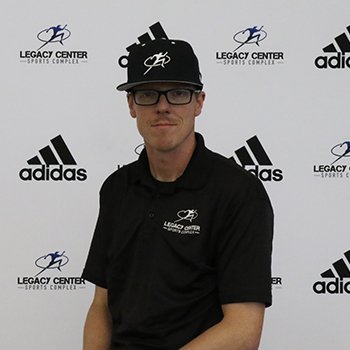 A young man wearing glasses and a black baseball cap with a logo, standing in front of a backdrop with various sports brand logos including Adidas and Legacy Center Sports Complex.