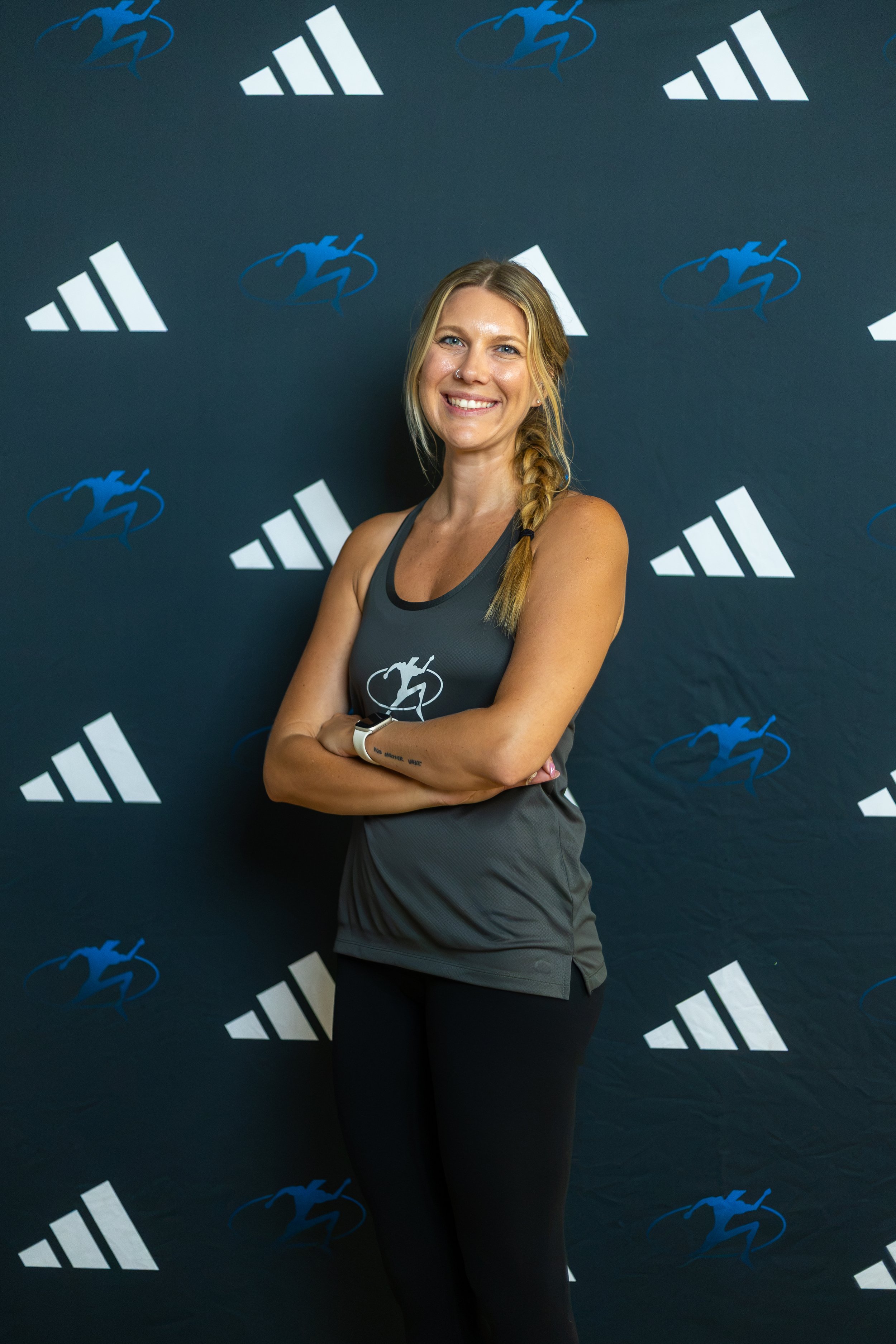 A woman with blonde hair tied back, wearing a black sleeveless top and a smartwatch, smiling and standing with arms crossed in a gym, leaning against a brown brick wall.