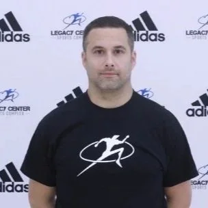 A man standing in front of a backdrop with Adidas and Legacy Center logos, wearing a black t-shirt with a white logo.