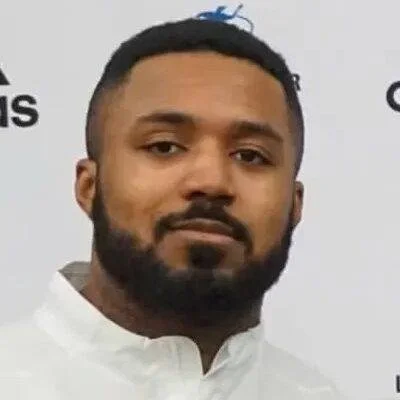A man with a beard and short hair wearing a white collared shirt, standing in front of a white background with partially visible logos.