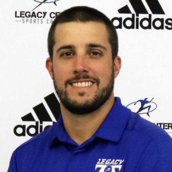 A smiling man with short dark hair and a beard, wearing a blue collared shirt with a logo, standing in front of a backdrop with Adidas logos and sports-related graphics.