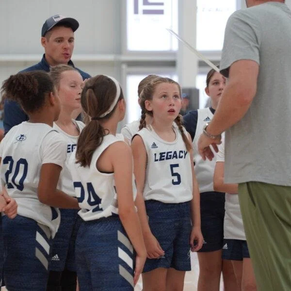 Young female volleyball players wearing white jerseys with blue shorts, listening to a coach with an orange clipboard, in an indoor gym.