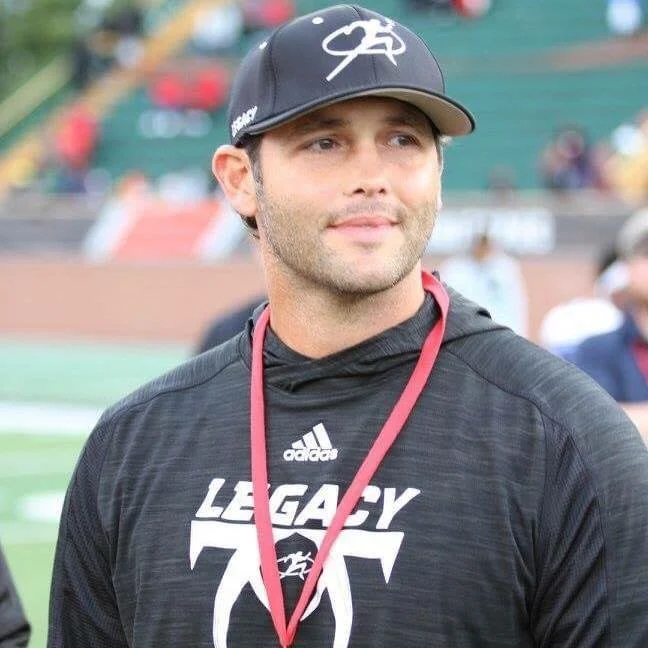 A man wearing a black Adidas hoodie with 'Legacy 70' logo, black cap with white logo, and a red medal lanyard around his neck, outdoors at a sports event.