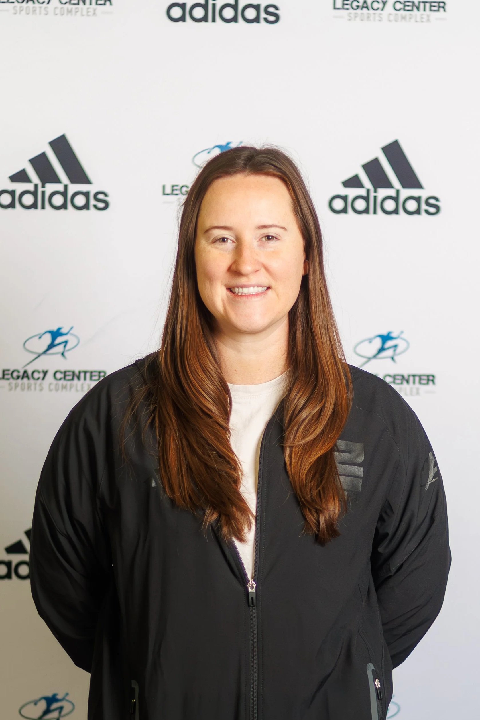 Young woman smiling in front of a backdrop with Adidas and Legacy Center logos, wearing a black sports jersey.