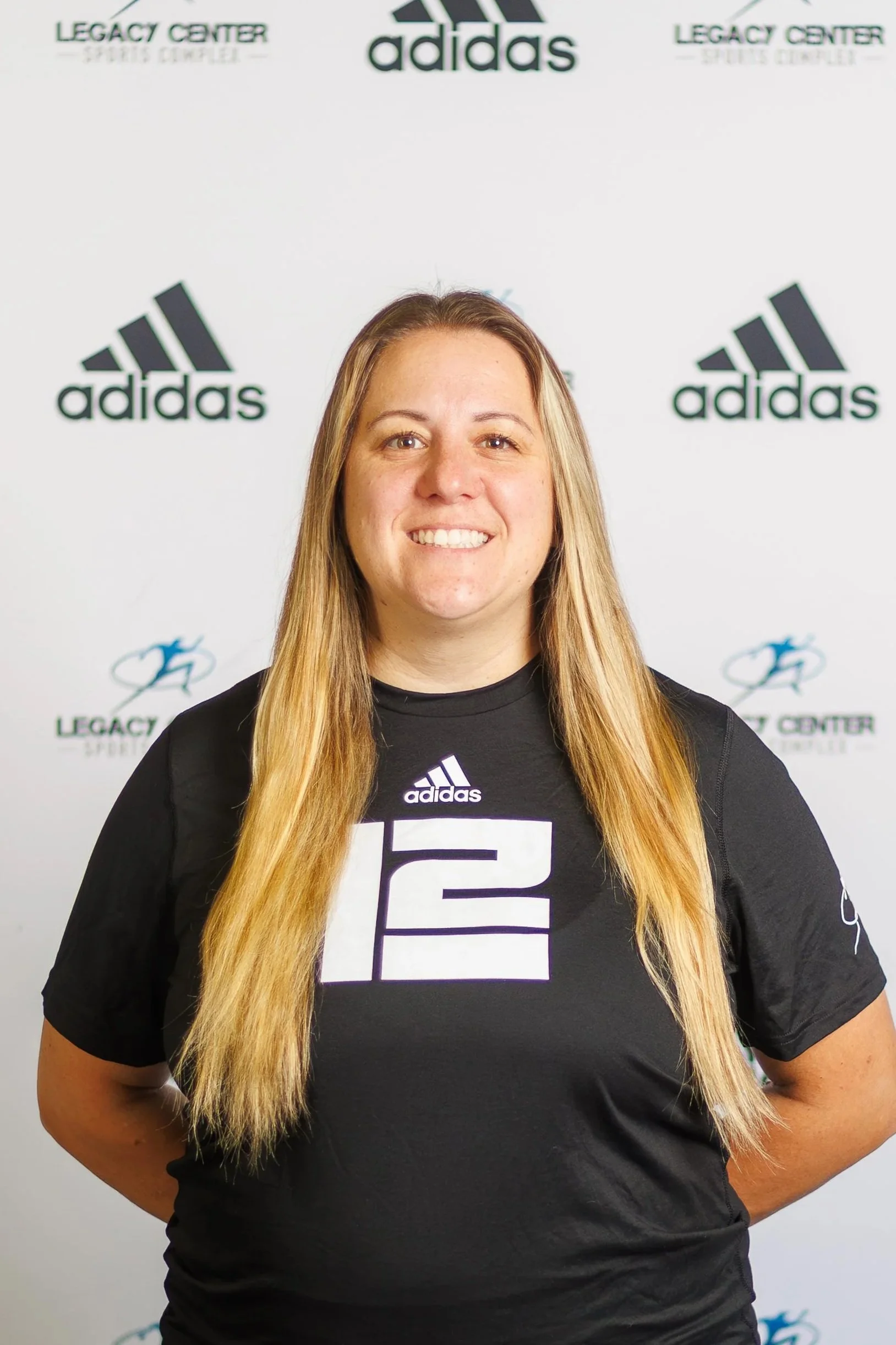 A smiling woman with long blonde hair wearing a black Adidas sports jersey standing in front of a backdrop with the Adidas logo, Legacy Center Sports Complex logo, and other sports-related graphics.