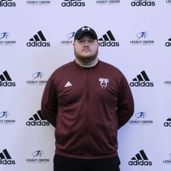 Man standing in front of a backdrop with Adidas and Legacy Center logos, wearing a maroon Athletic jacket, black baseball cap, and with hands behind back.