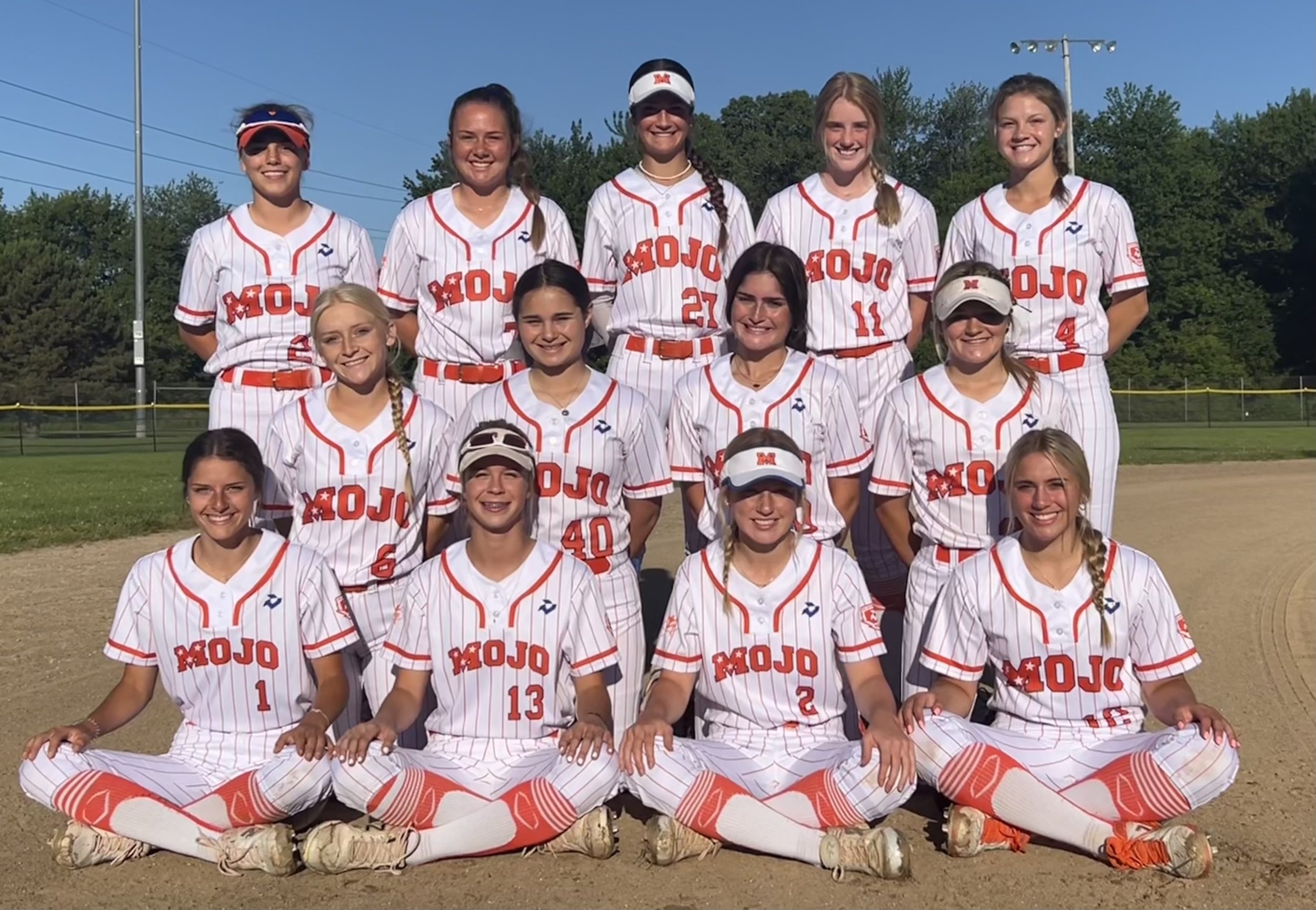 Group of young female softball players in white pinstripe uniforms with red lettering, standing on a baseball field on a sunny day.