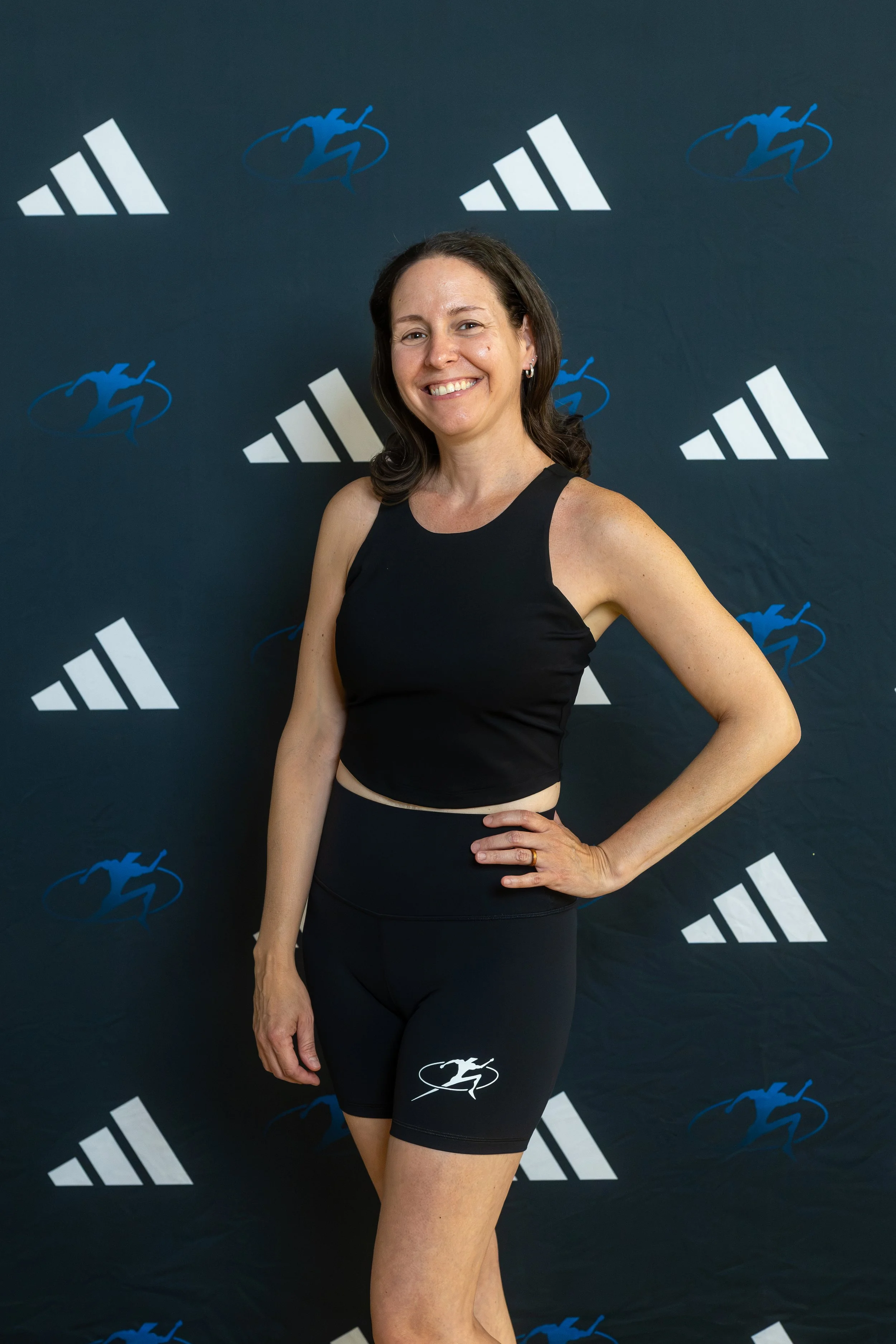 A woman with dark brown hair smiling in a gym, wearing a black Adidas long-sleeve shirt, with gym equipment and flags hanging from the ceiling in the background.