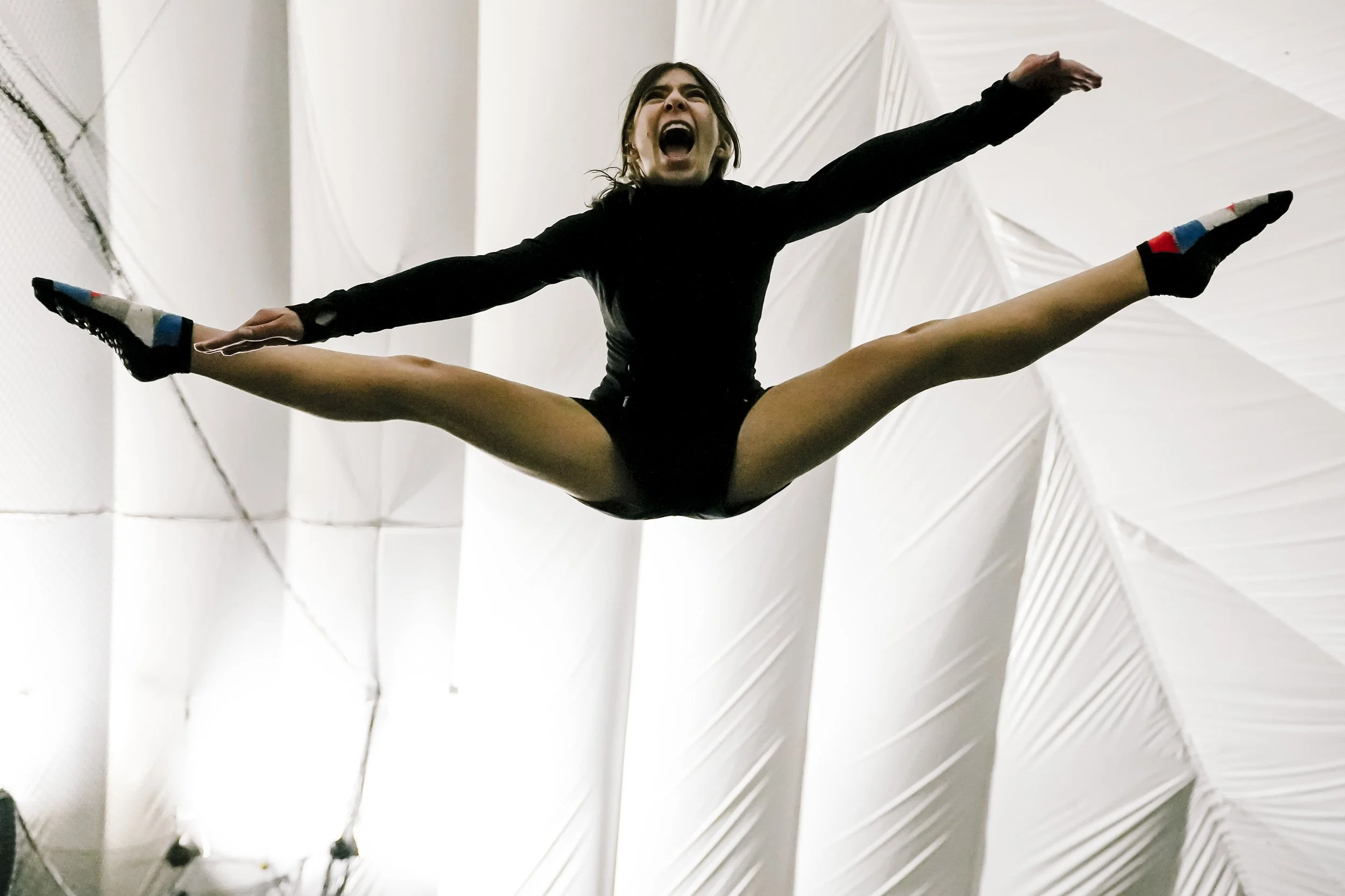 A female gymnast performing a split leap in mid-air with arms outstretched and mouth open in an expression of effort or excitement, wearing a black leotard and colorful socks, against a white indoor sports facility ceiling.