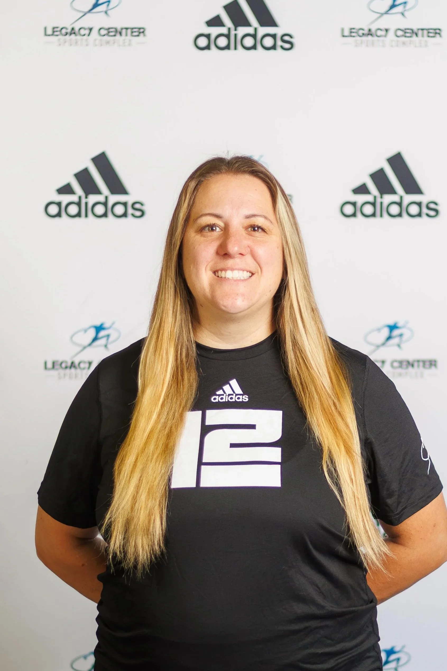 A young woman with long blonde hair smiling, standing in front of a backdrop with Adidas and Legacy Center logos.
