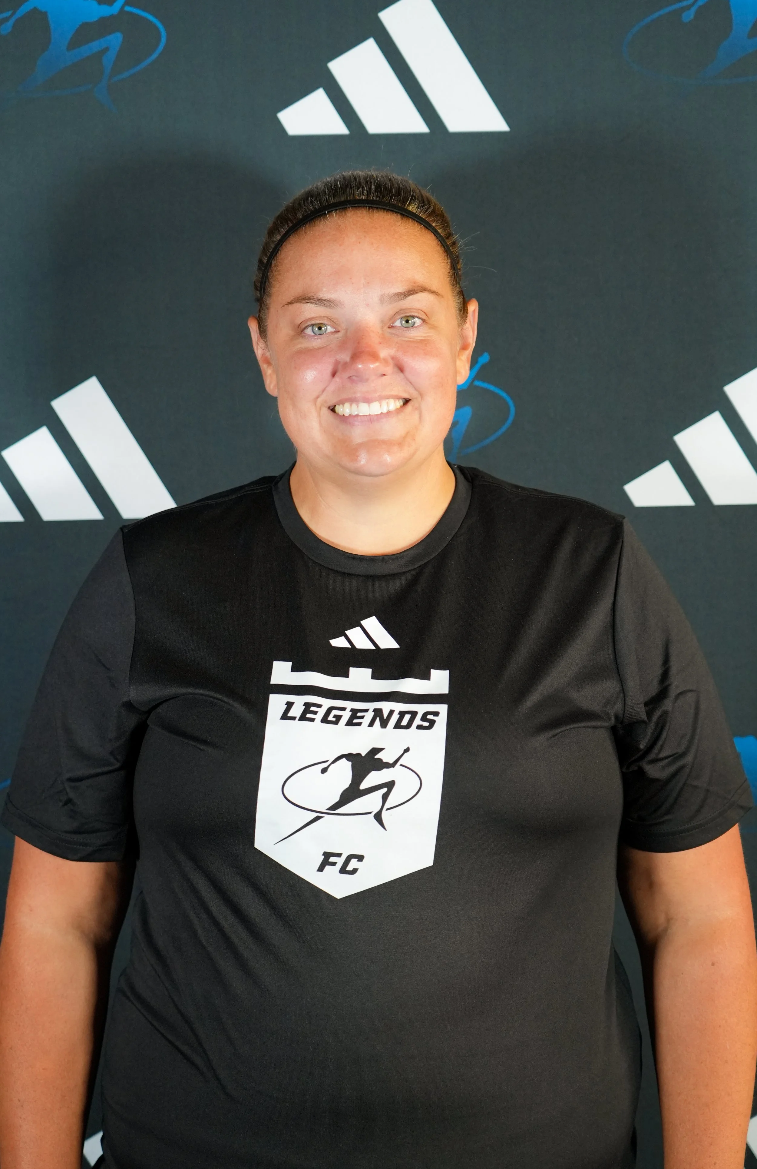 Young woman wearing a black T-shirt with gold text, a black headband, and smiling, standing in front of a white background with logos of Legacy Center Sports Complex and Adidas.