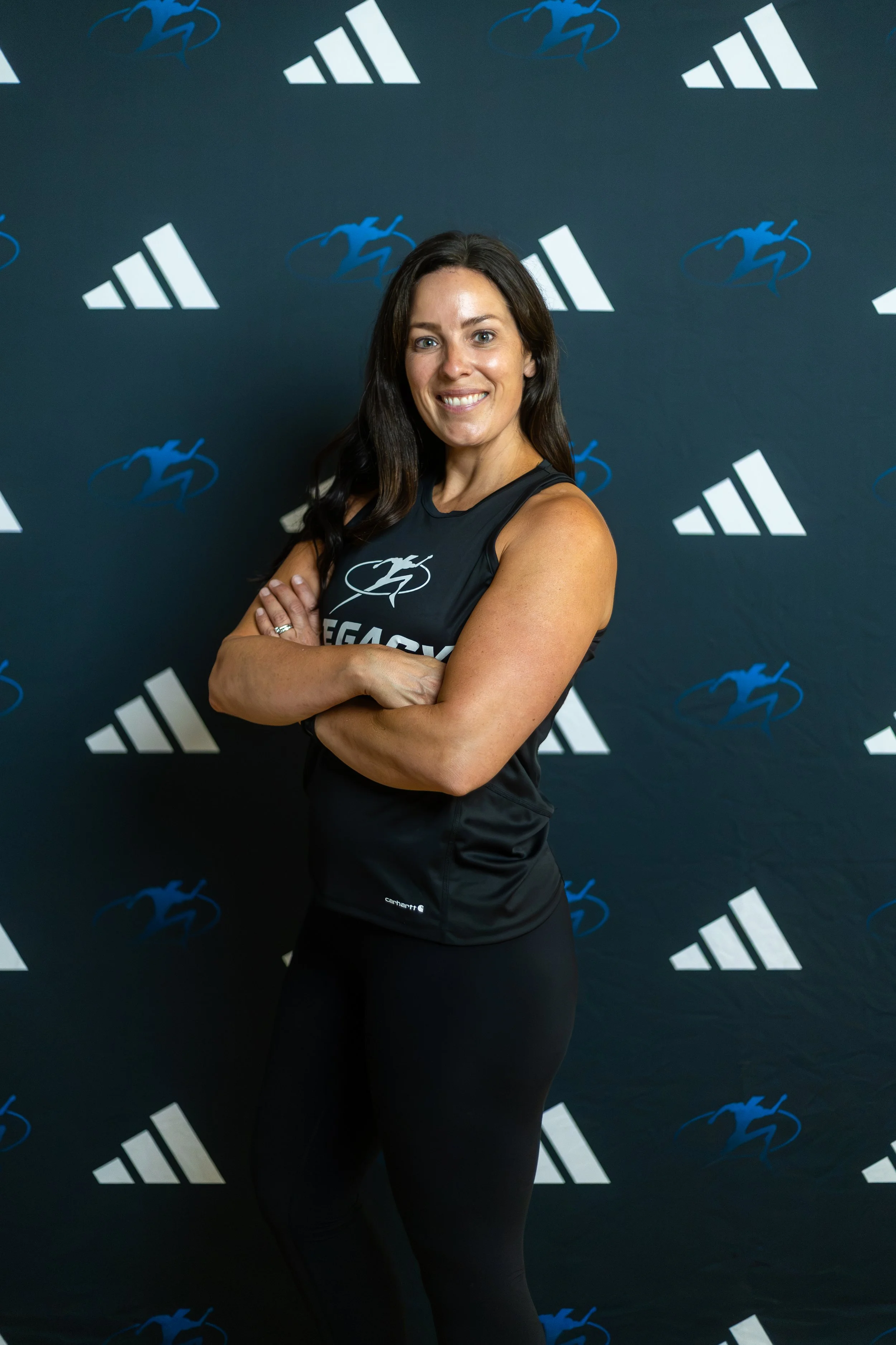 A smiling woman with dark hair in a gym, standing with arms crossed beside exercise equipment.