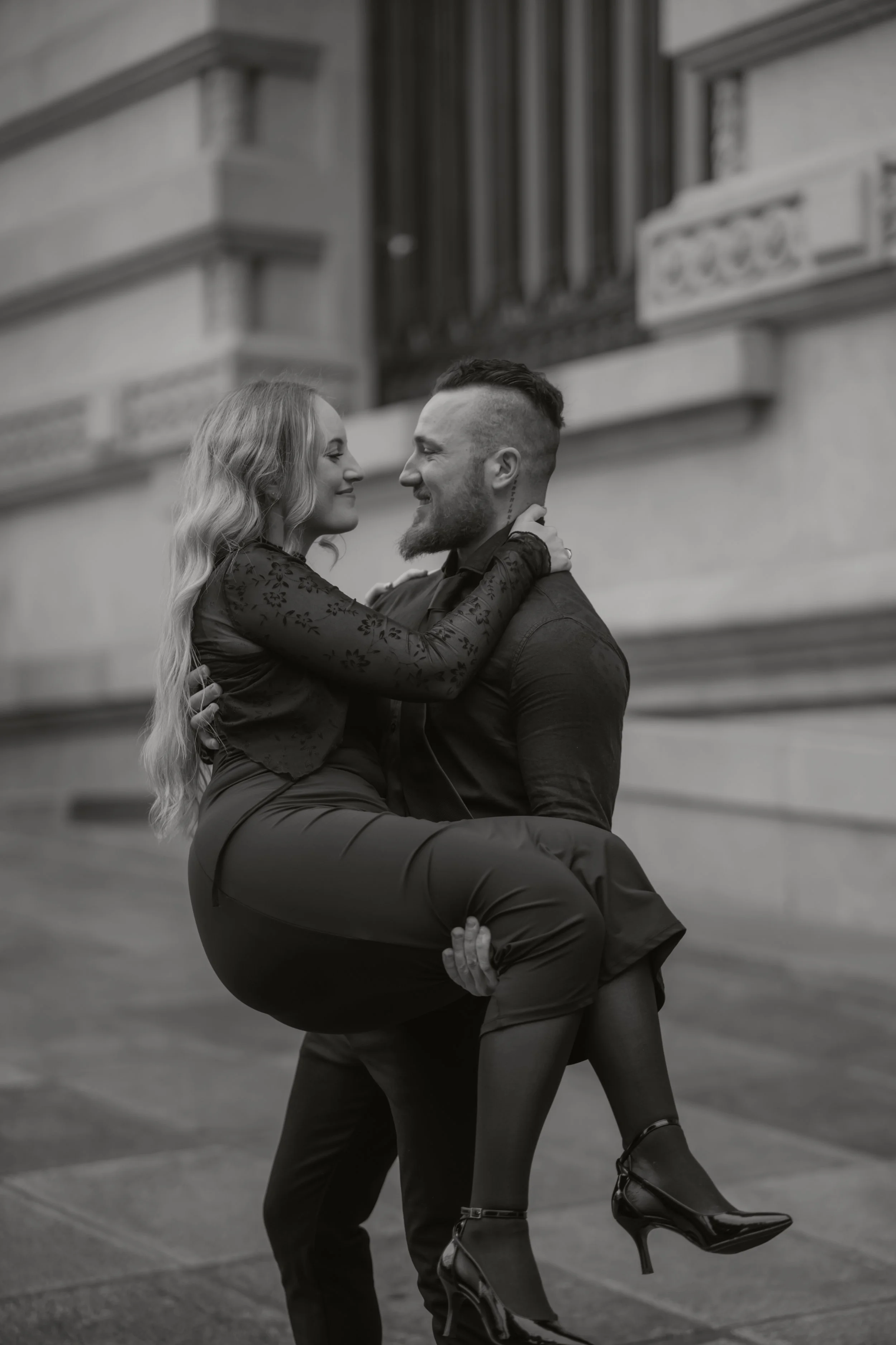 Ein Mann trägt eine Frau, beide lächeln sich an, im schwarzen und weißen Foto vor einer alten Gebäudefassade.