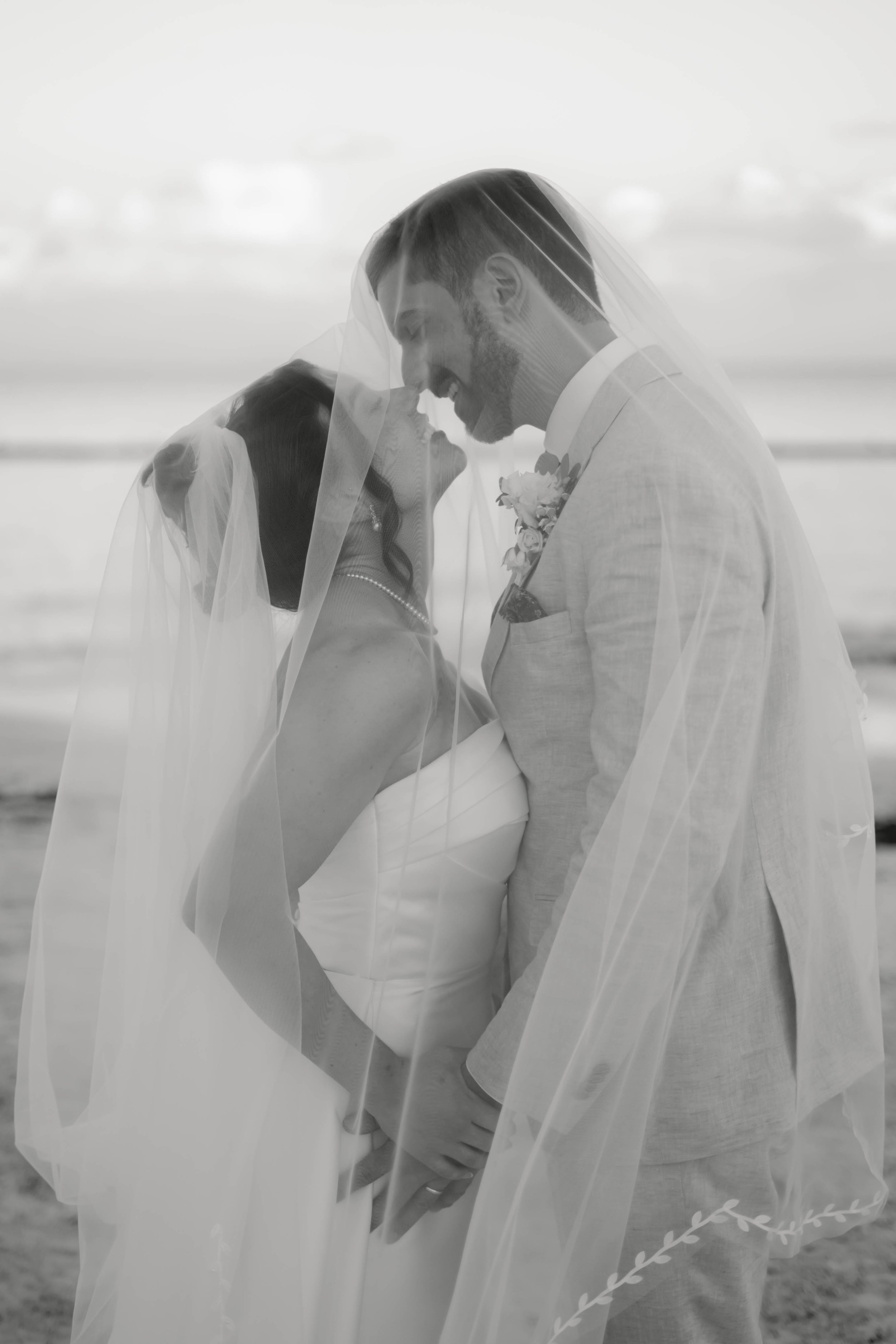 Braut und Bräutigam unter einem Schleier, lächelnd, am Strand, in Schwarz-Weiß, bei einer Hochzeit.