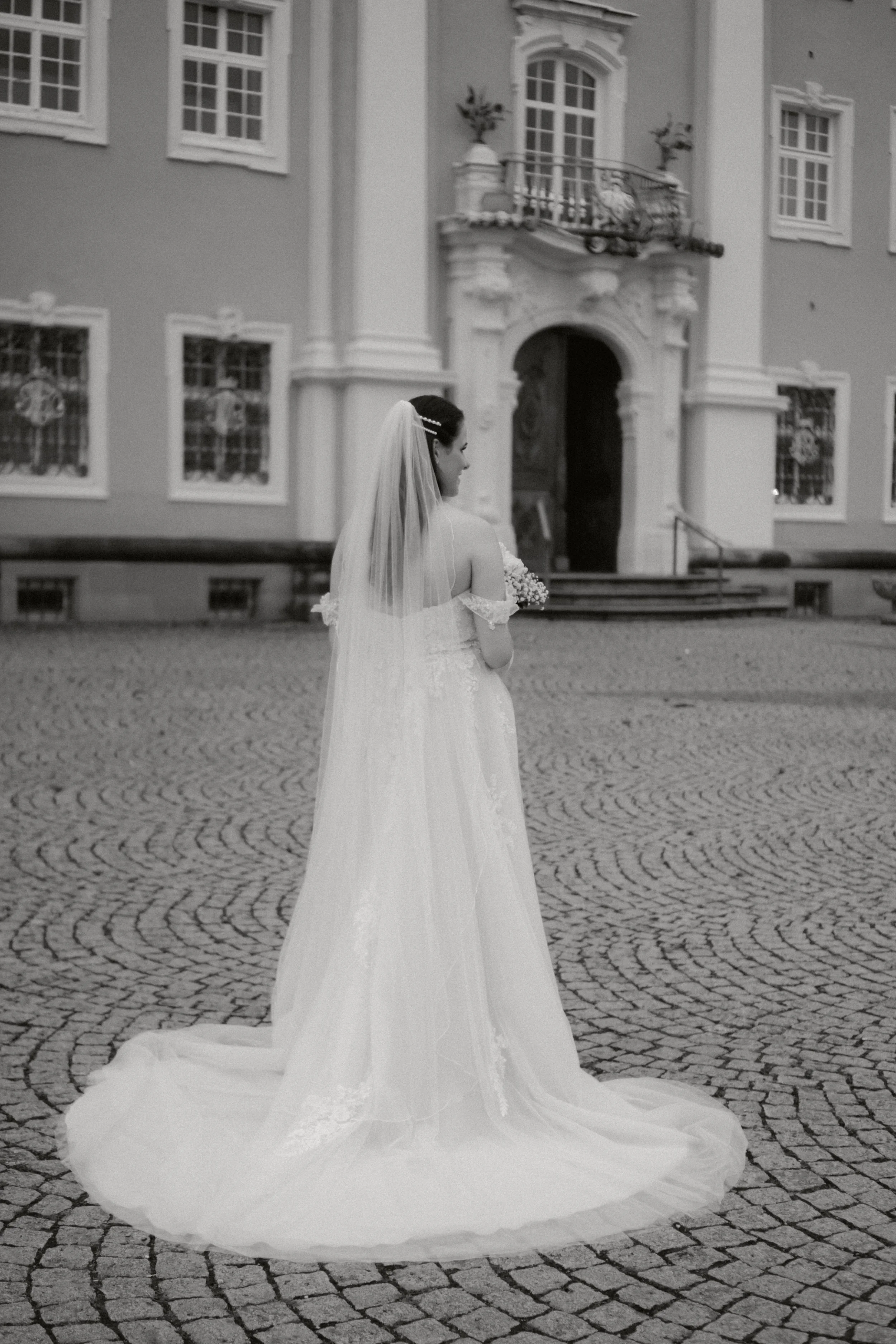 Braut in Hochzeitskleid mit Schleier vor einem alten, barocken Gebäude auf gepflastertem Platz.