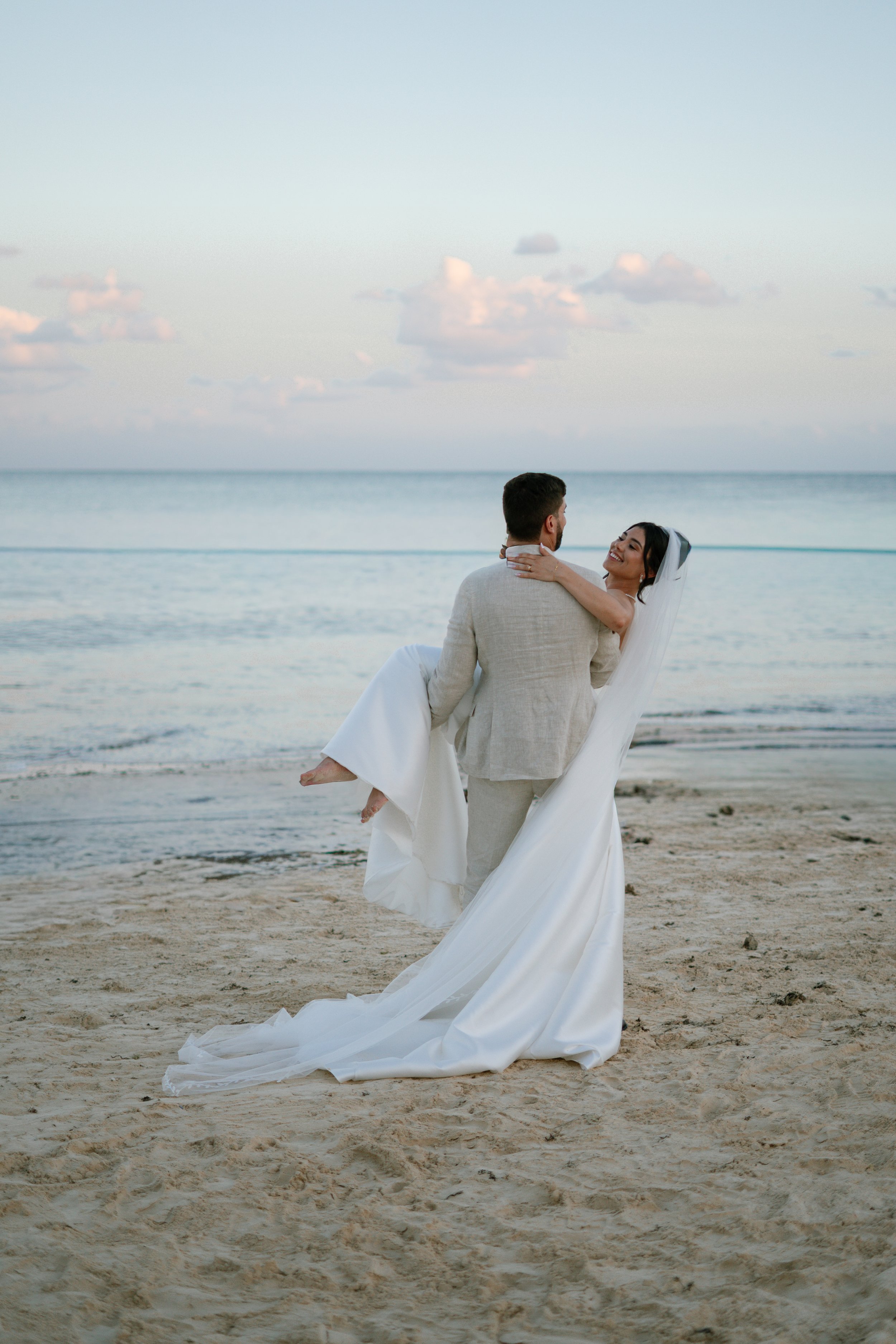 Ein Hochzeitsfoto am Strand mit einem Mann, der eine Frau trägt, beide in Hochzeitskleidung, bei Sonnenuntergang.