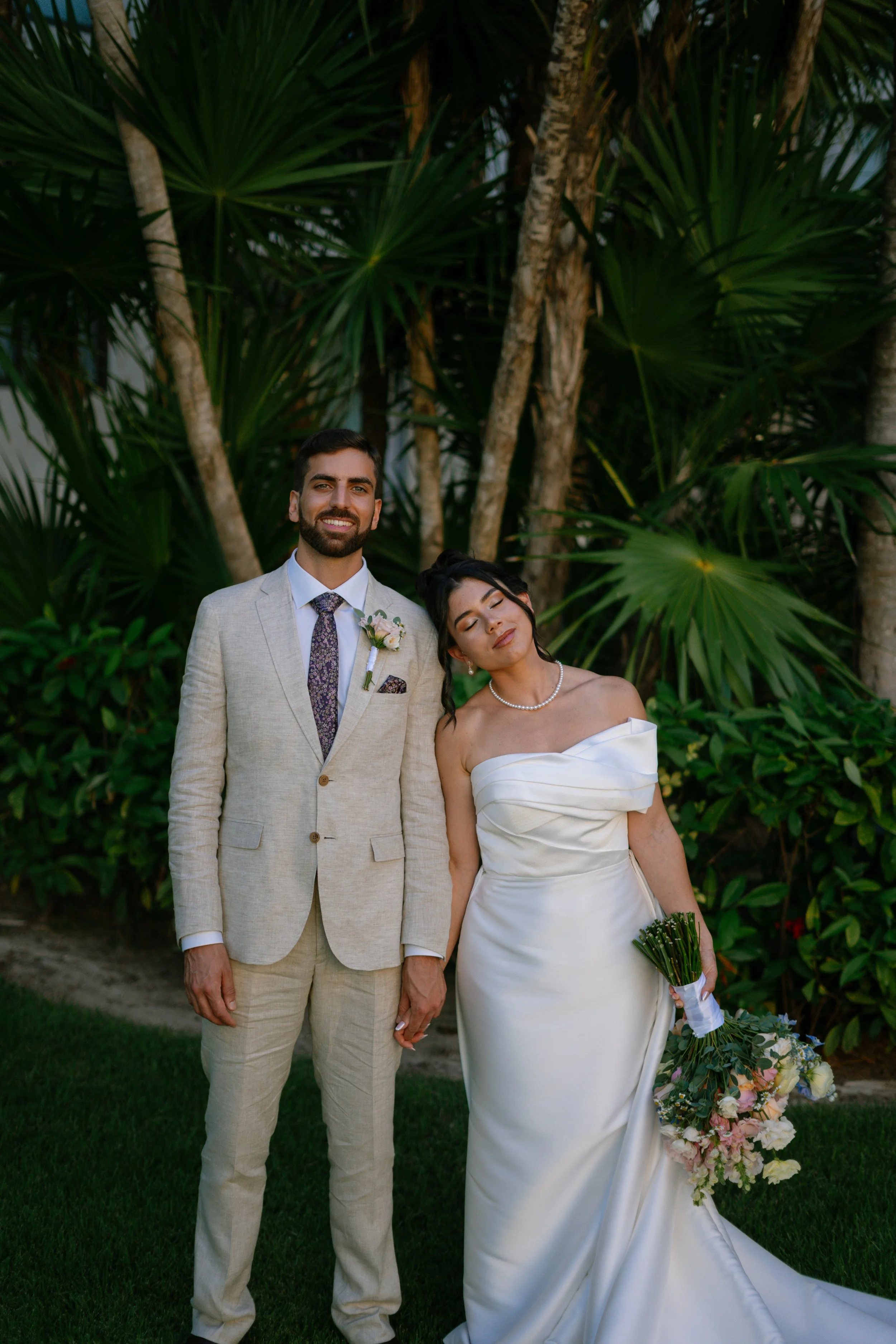 Ein Brautpaar steht im Freien vor grünen Palmen. Der Bräutigam trägt einen hellgrauen Anzug mit Krawatte, die Braut trägt ein weißes, schulterfreies Hochzeitskleid und hält einen Blumenstrauß.